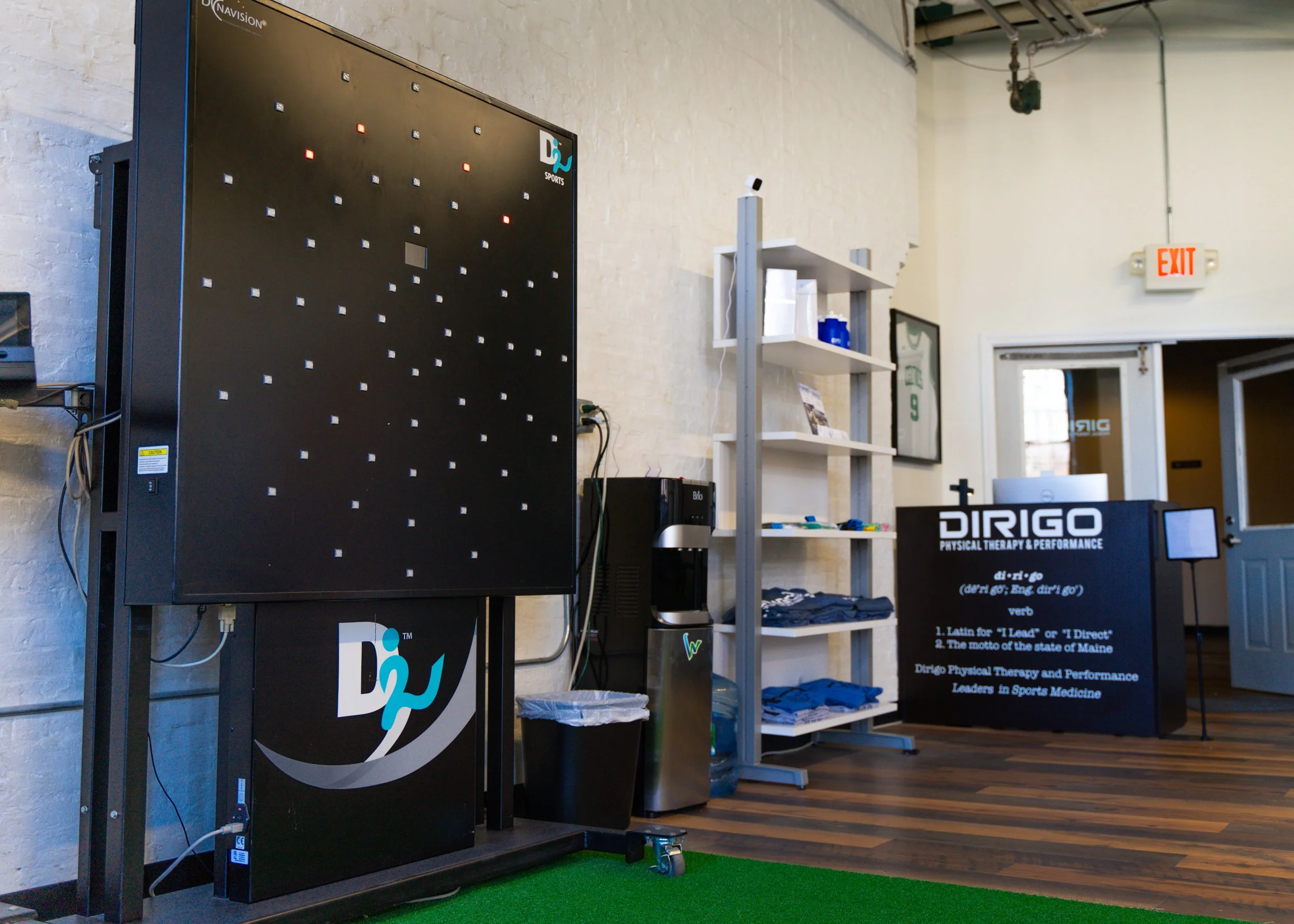Interior of Dirigo Physical Therapy & Performance showing a training wall with lights, shelves with equipment and apparel, a water cooler, and a reception desk.