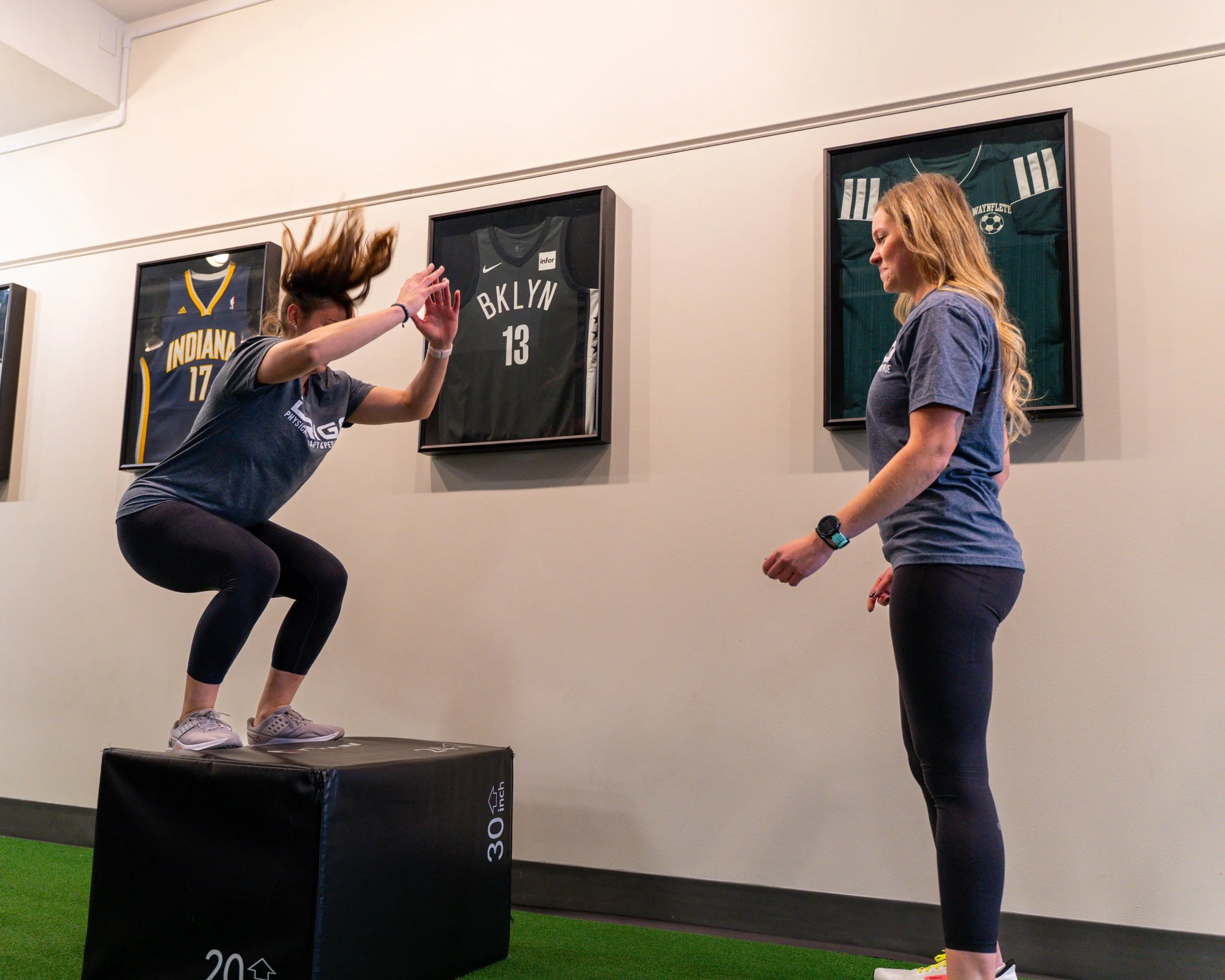 Two women in a gym, one jumping on a soft box while the other observes, framed jerseys on the wall