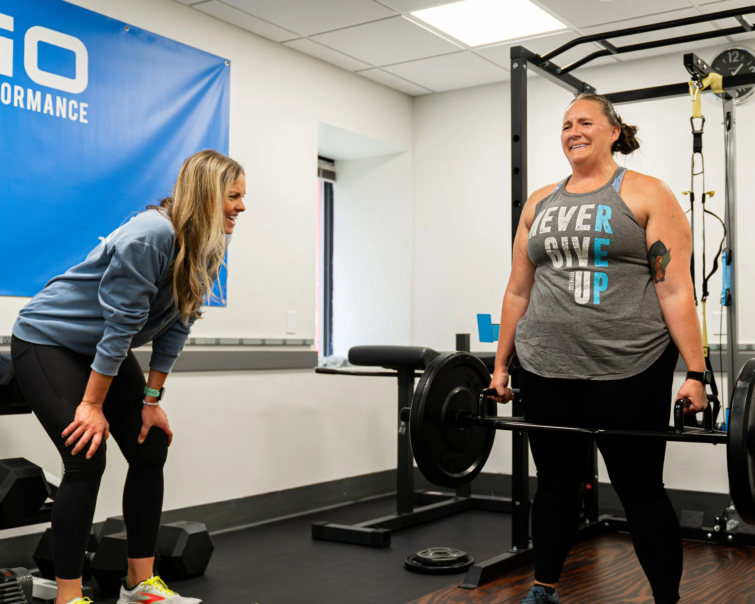 Two people exercising indoors, one holding dumbbells, with framed sports jerseys on the wall in the background.