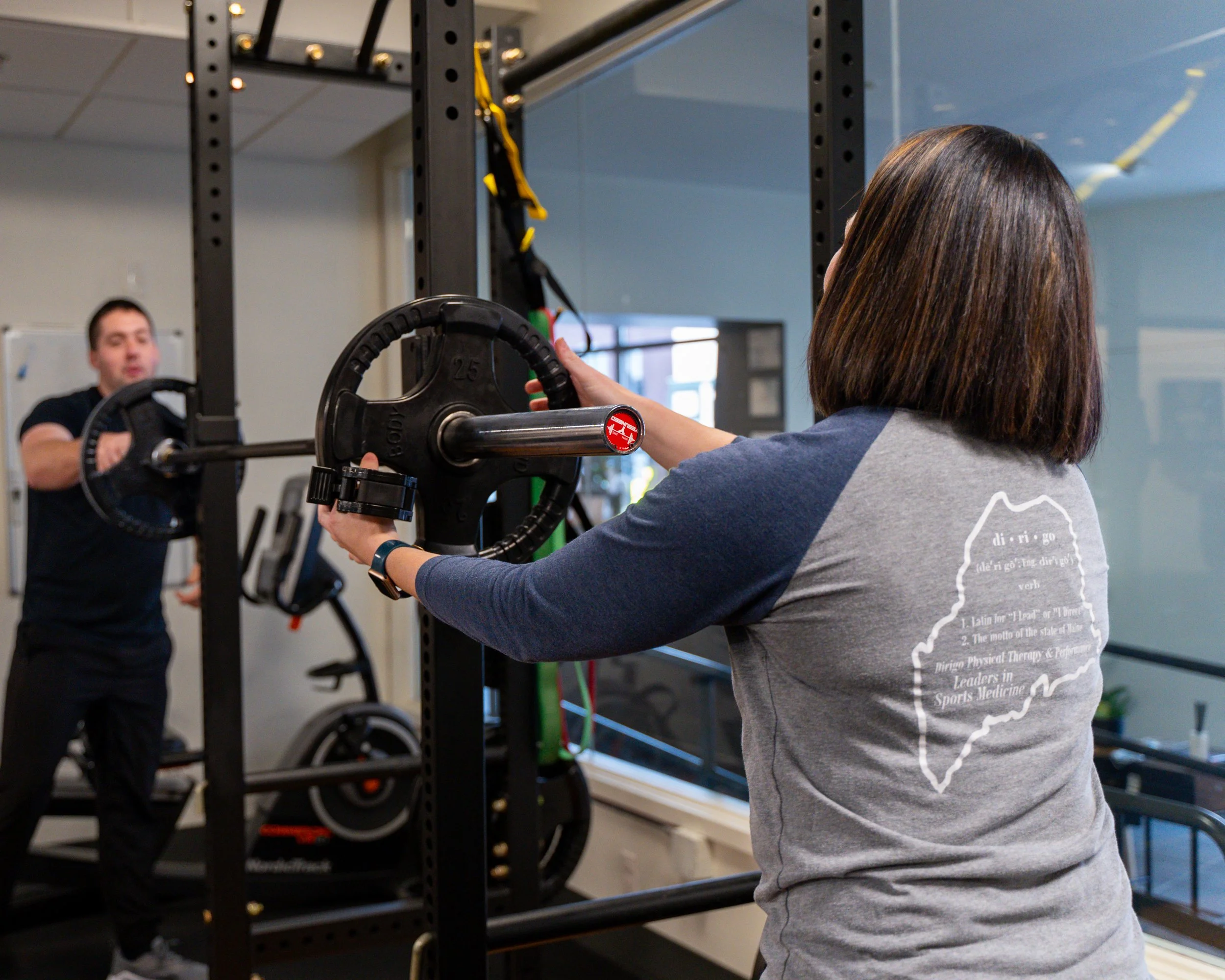 Two people in a gym setting with one attaching a weight plate to a barbell on a squat rack, while the other appears to assist or observe.