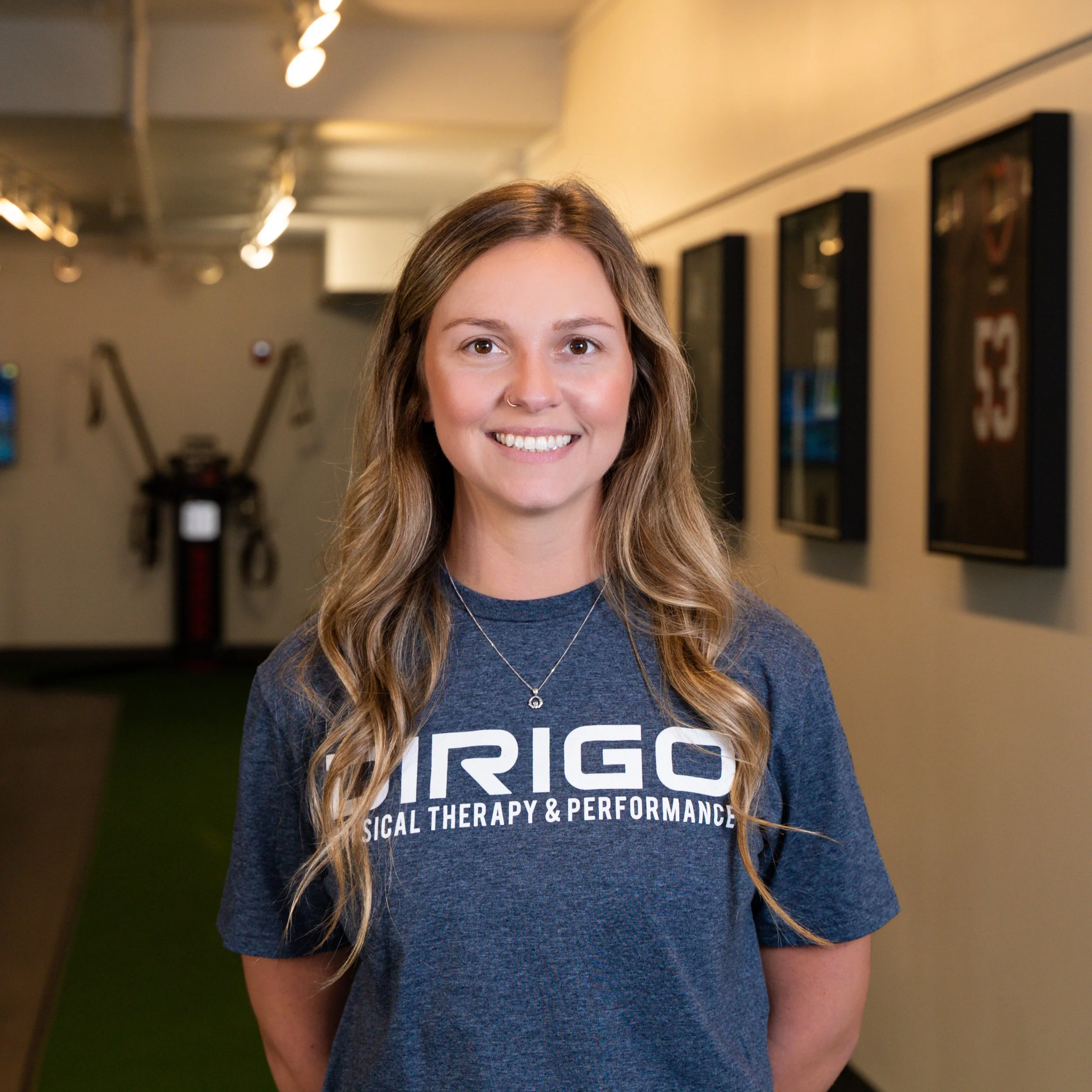 Person smiling in a Dirigo Physical Therapy & Performance t-shirt, standing in a hallway with sports memorabilia on the walls.