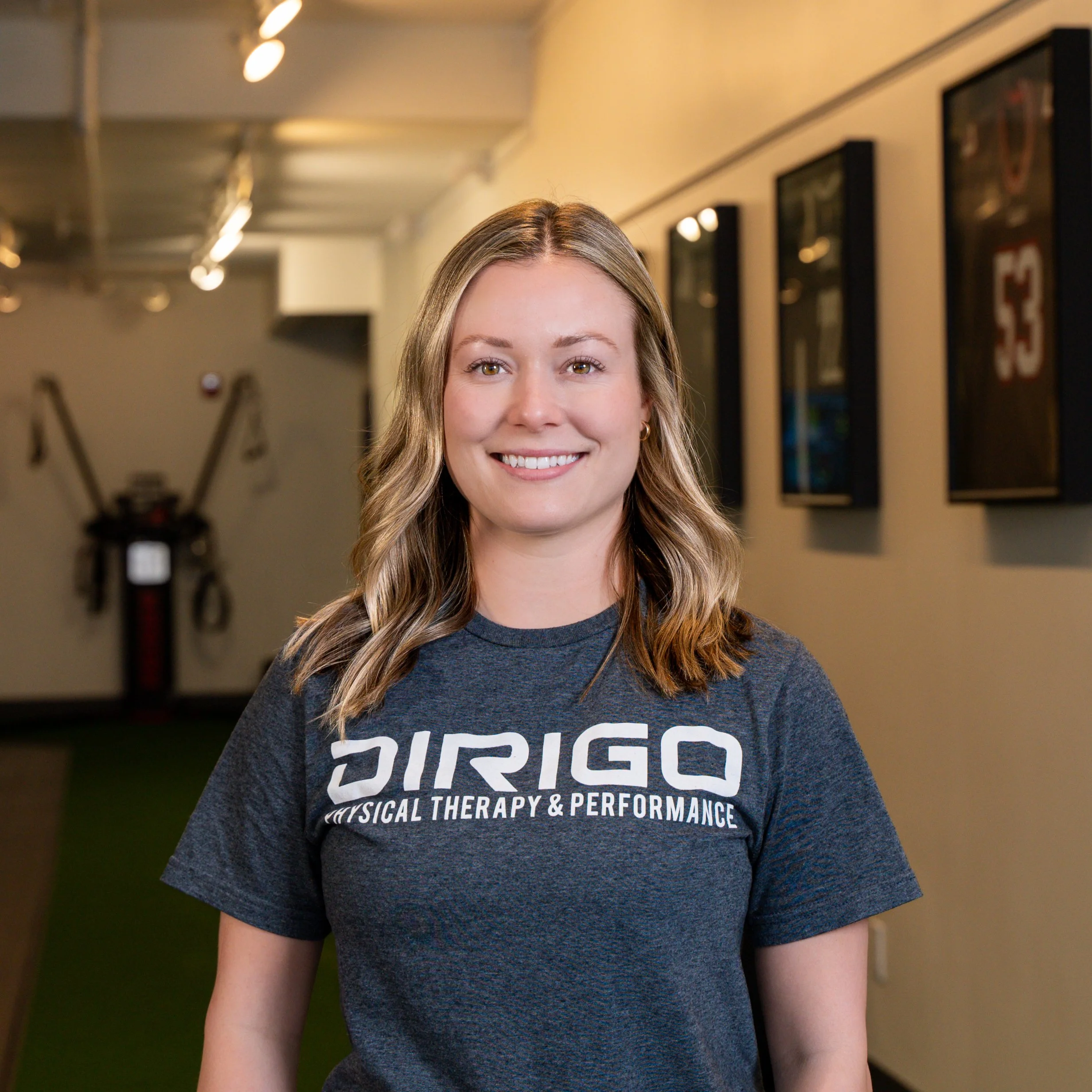 Woman smiling inside a physical therapy clinic, wearing a shirt with the text "DIRIGO PHYSICAL THERAPY & PERFORMANCE."