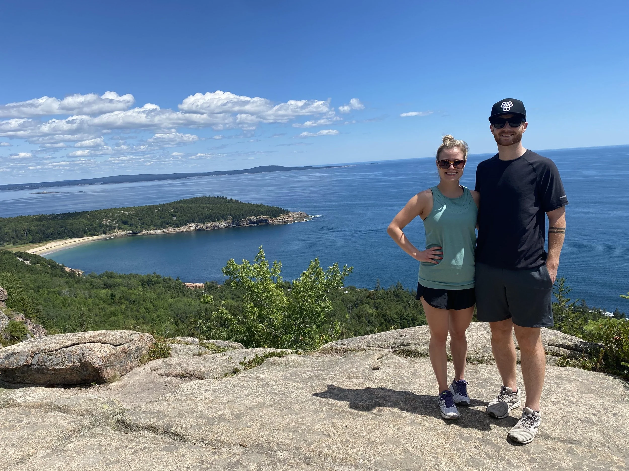 A couple stands on a rocky ledge overlooking a coastal landscape with lush greenery, sandy beach, and blue ocean under a clear, cloudy sky.