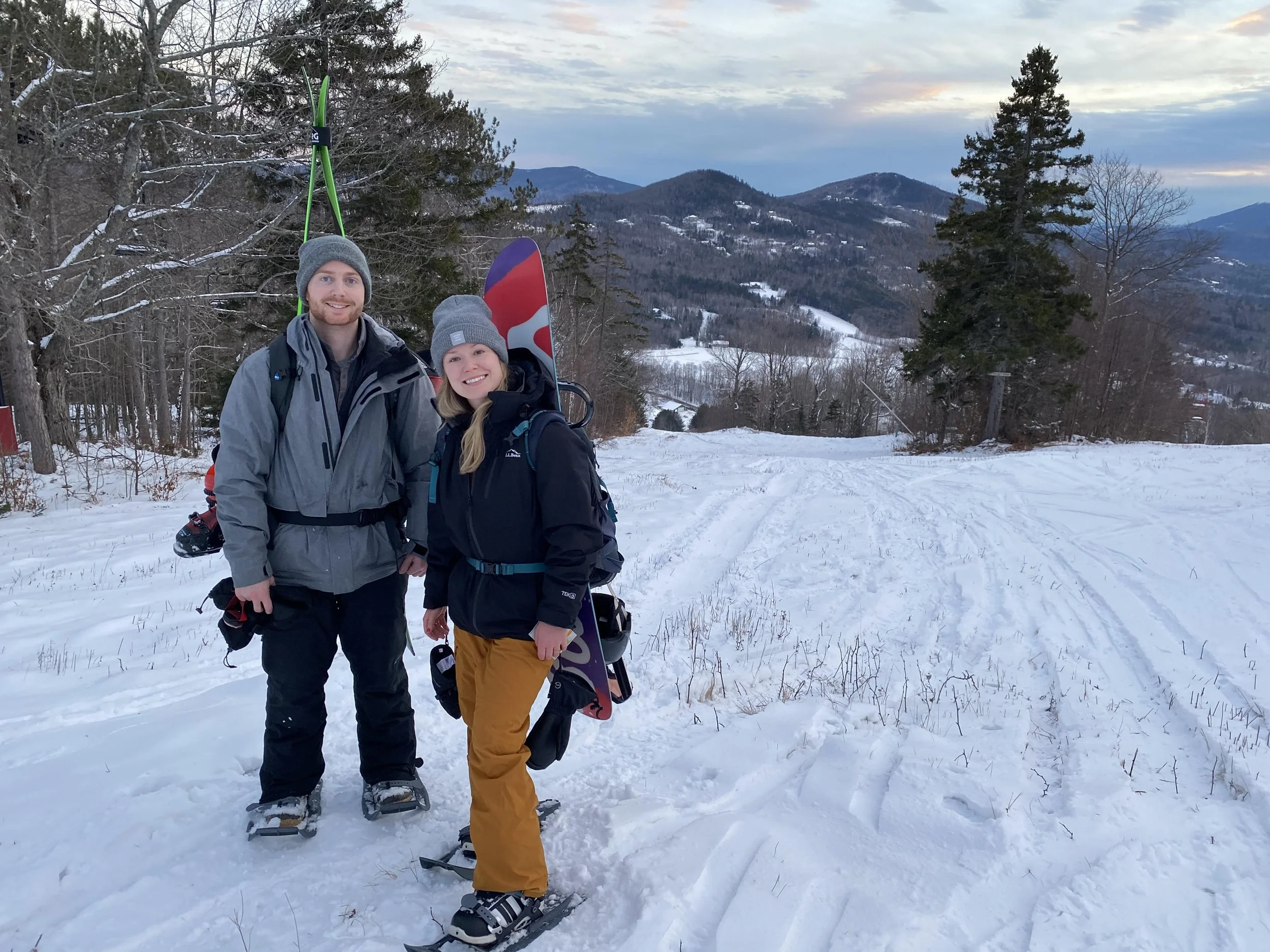 Two people in winter clothing and snowshoes standing in a snowy landscape with mountains and trees in the background, carrying snowboards.