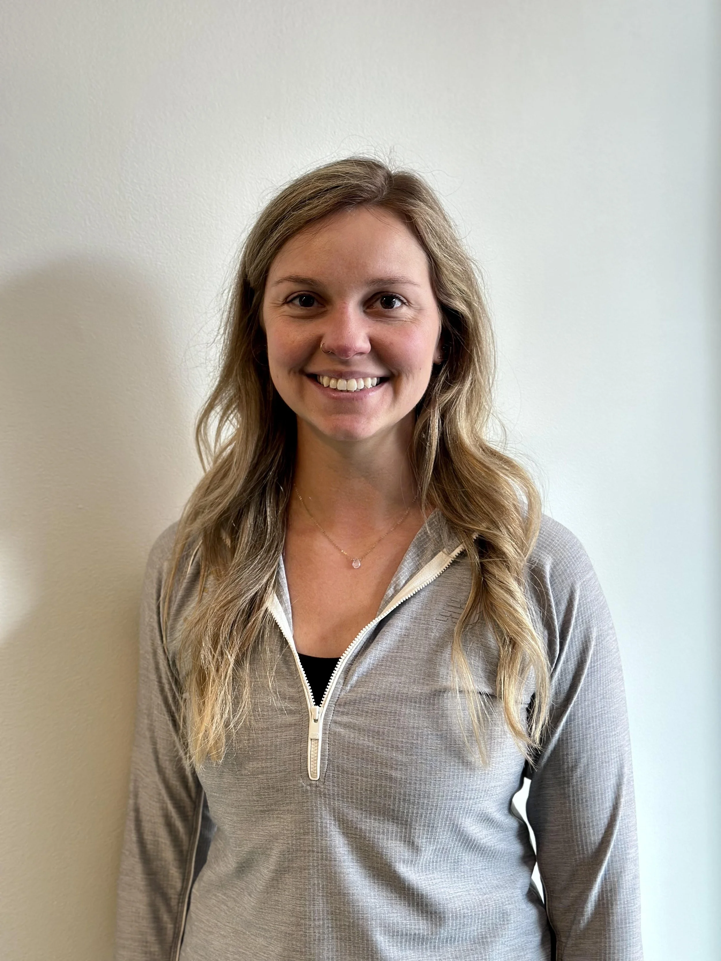 A woman with long blonde hair wearing a gray zipper-up top standing against a plain white background, smiling at the camera.