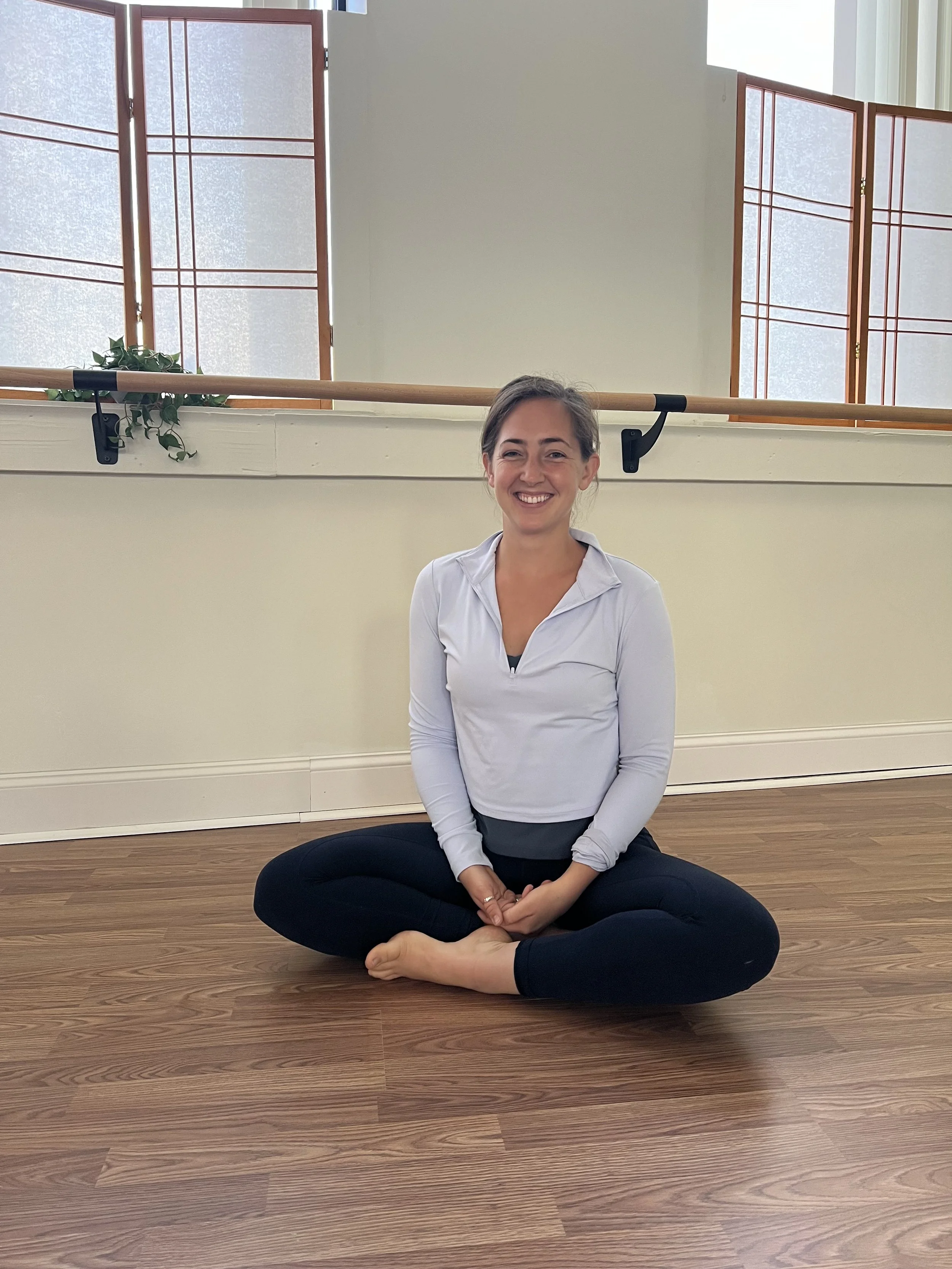 Woman sitting cross-legged on wooden floor, smiling, in a room with ballet barre and windows.