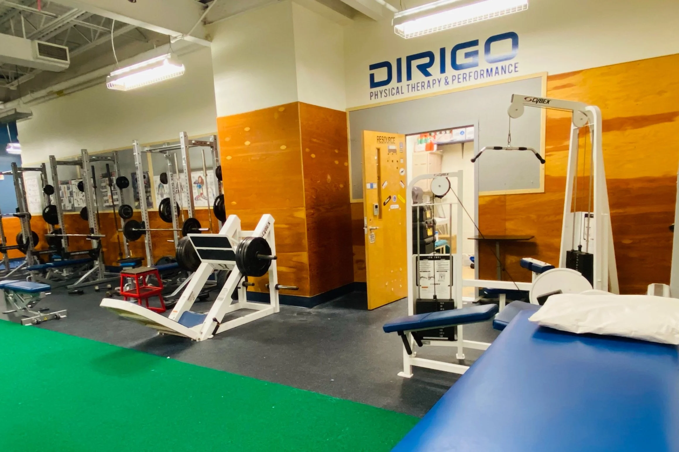 Physical therapy and performance gym with exercise equipment, including weight racks, a leg press machine, and a therapy table, inside a room labeled "Dirigo Physical Therapy & Performance."