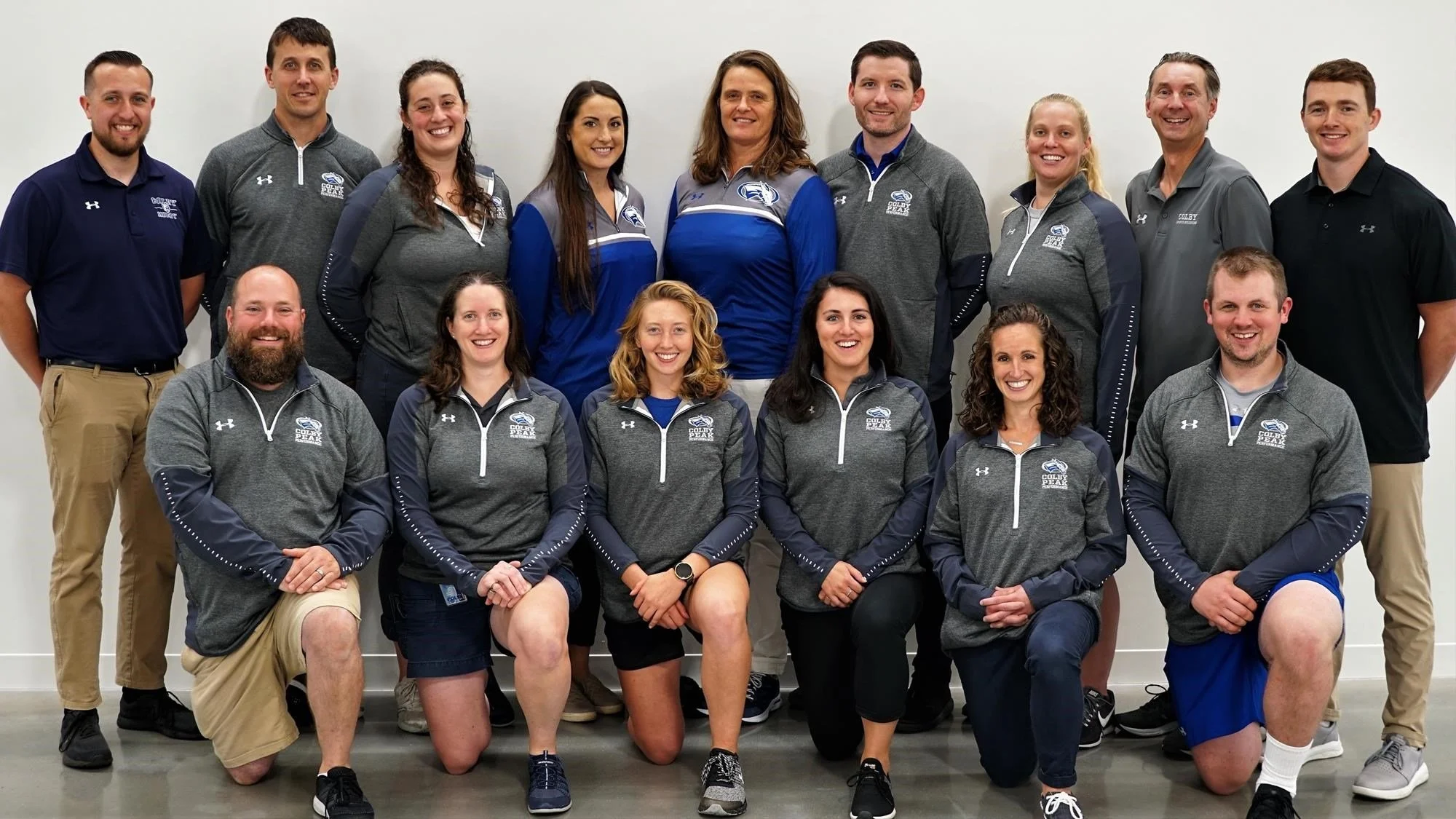 Group photo of sports team staff wearing matching grey and blue uniforms, posed against a plain white background.