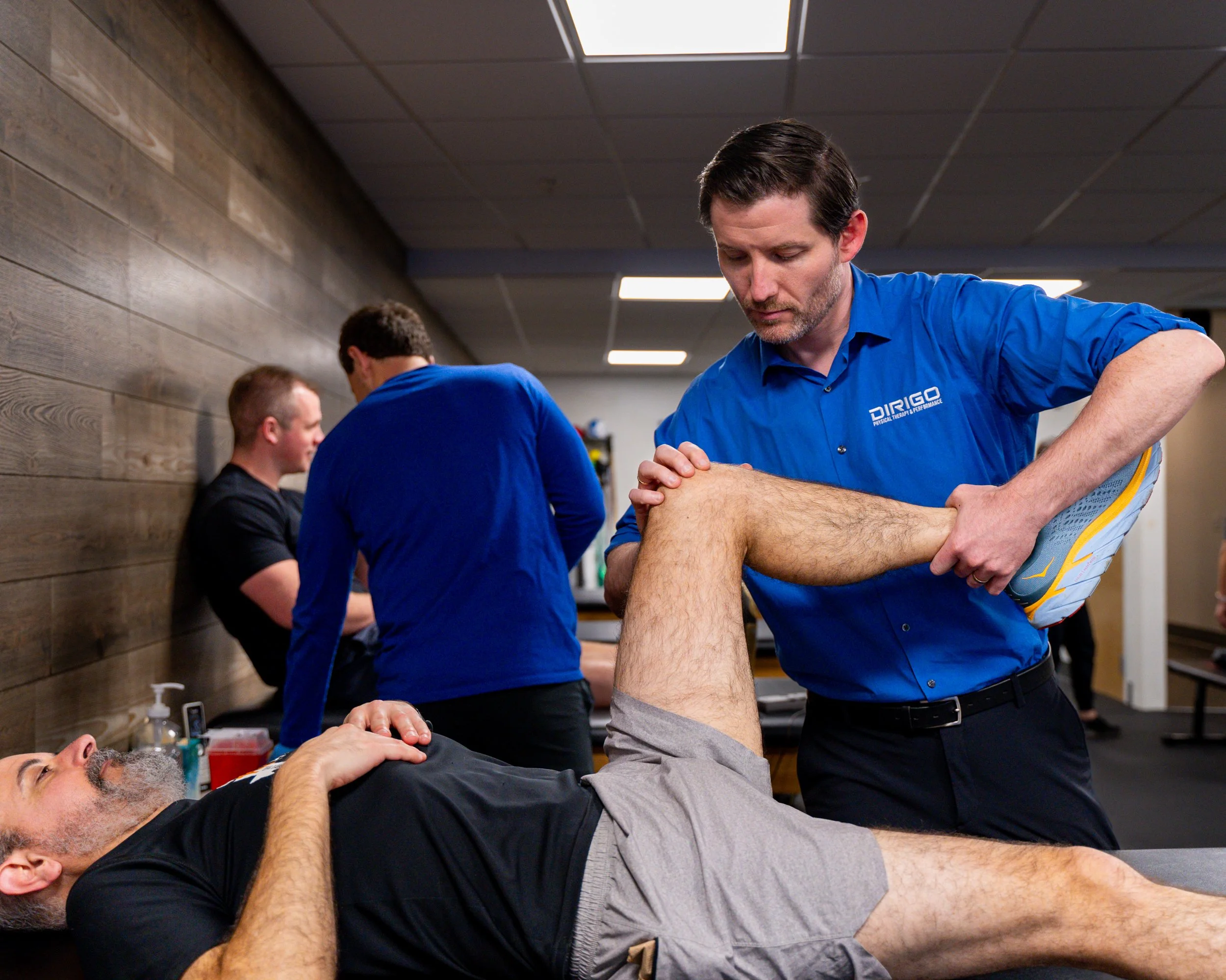 A physical therapist in a blue shirt performing leg exercises on a patient lying on a table in a clinical setting.