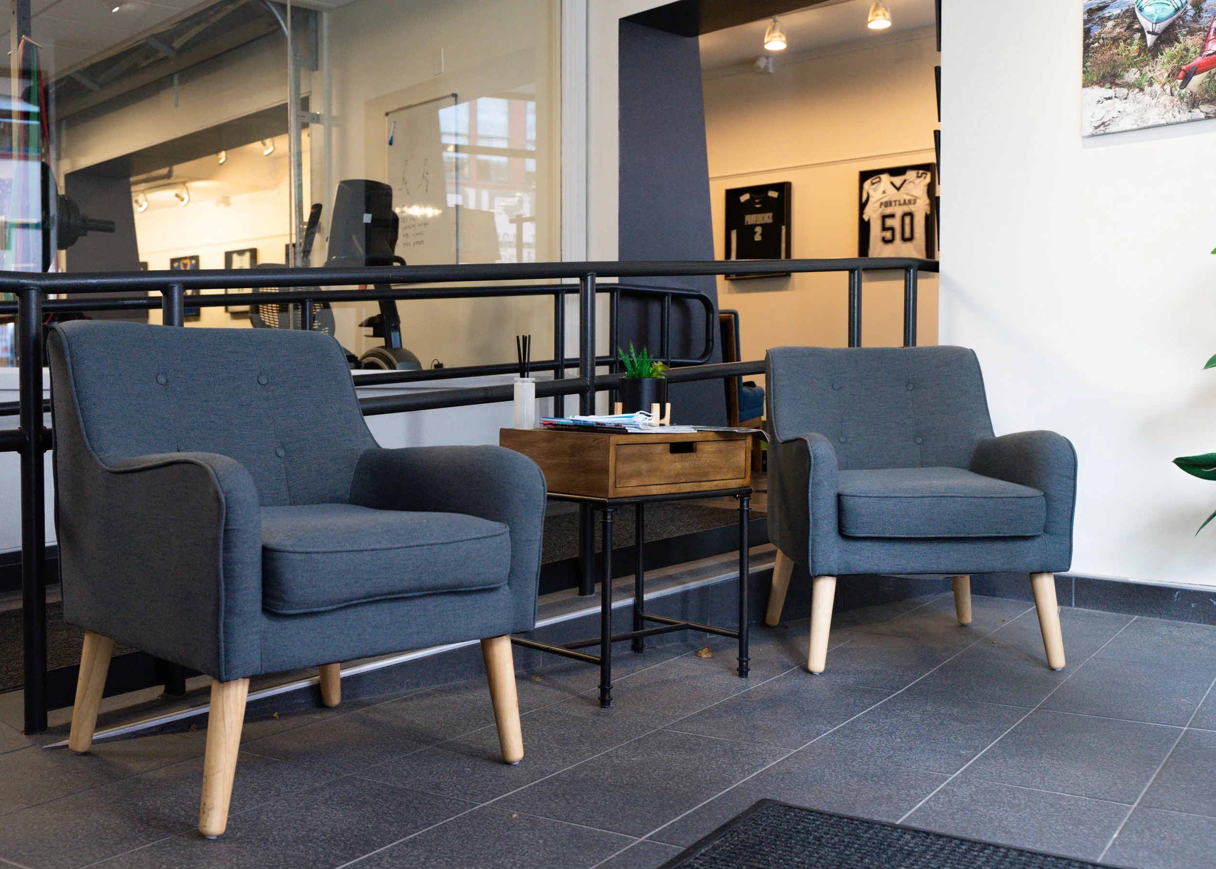 Two gray armchairs with wooden legs flanking a wooden side table in a modern room with sports decor on the walls.