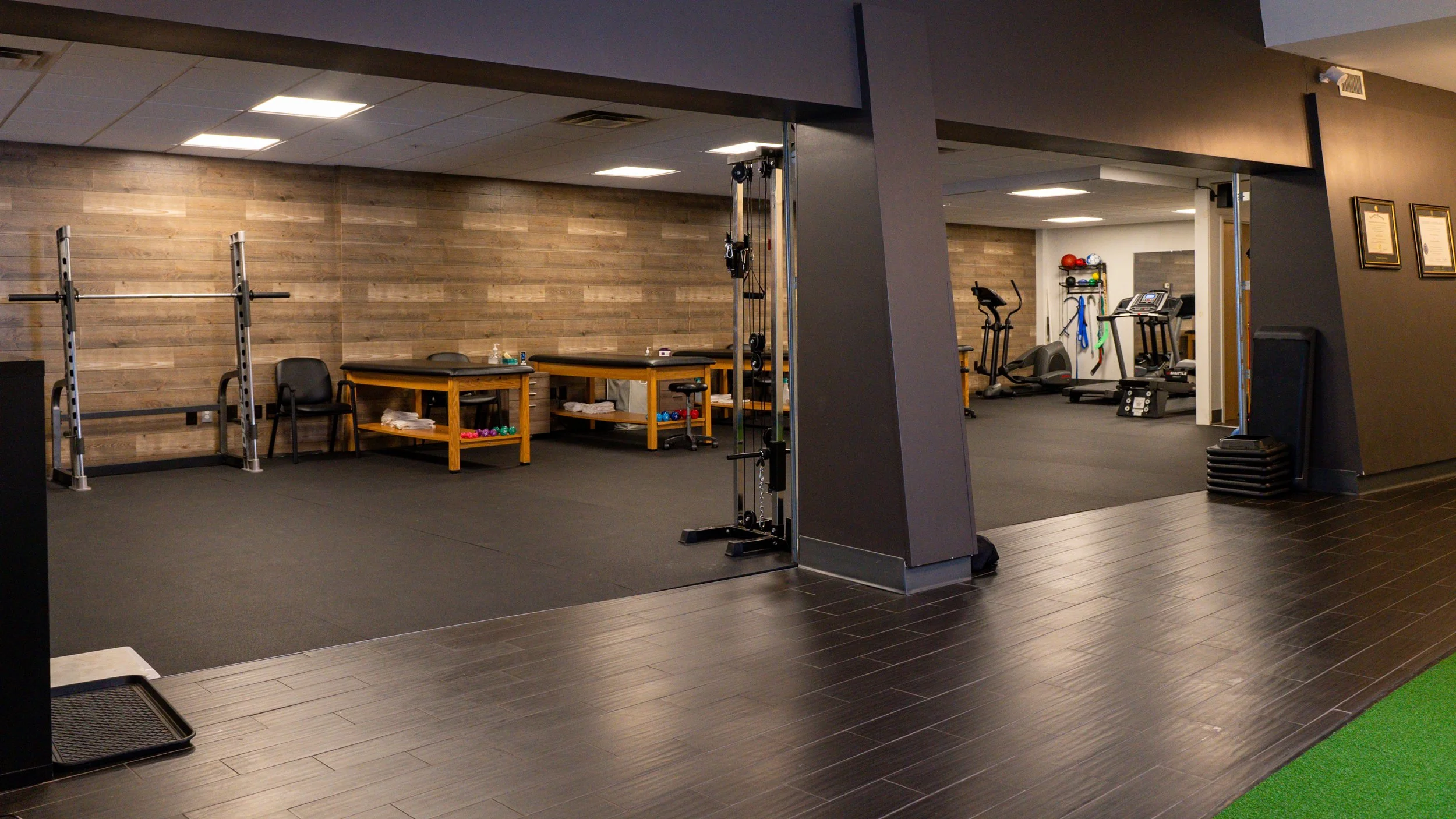 Interior of a fitness center with exercise equipment, including a squat rack, weight benches, free weights, stationary bikes, and treadmills, set against wood-paneled walls.