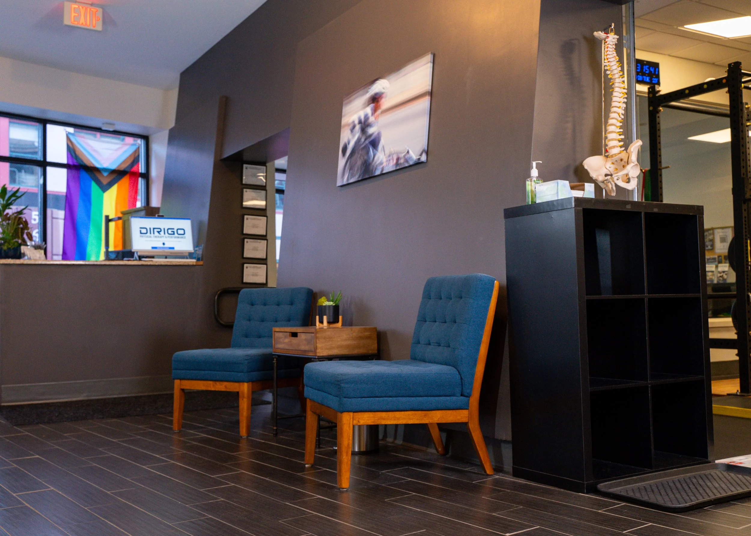 Modern waiting room with blue chairs, small table, spinal column model, and rainbow flag in the background.