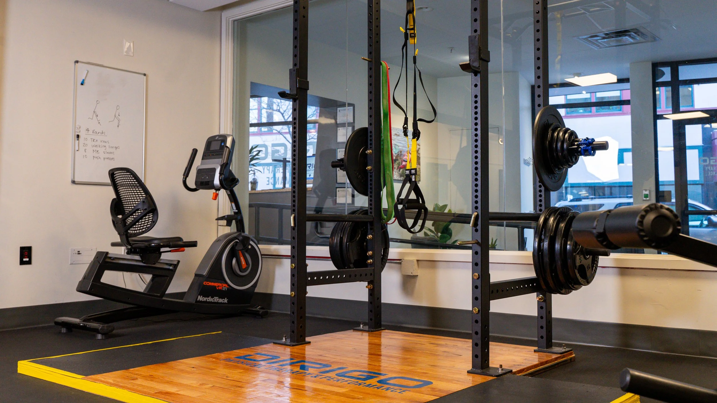 Gym interior with exercise bike, squat rack, weights, TRX bands, and a whiteboard with workout instructions.