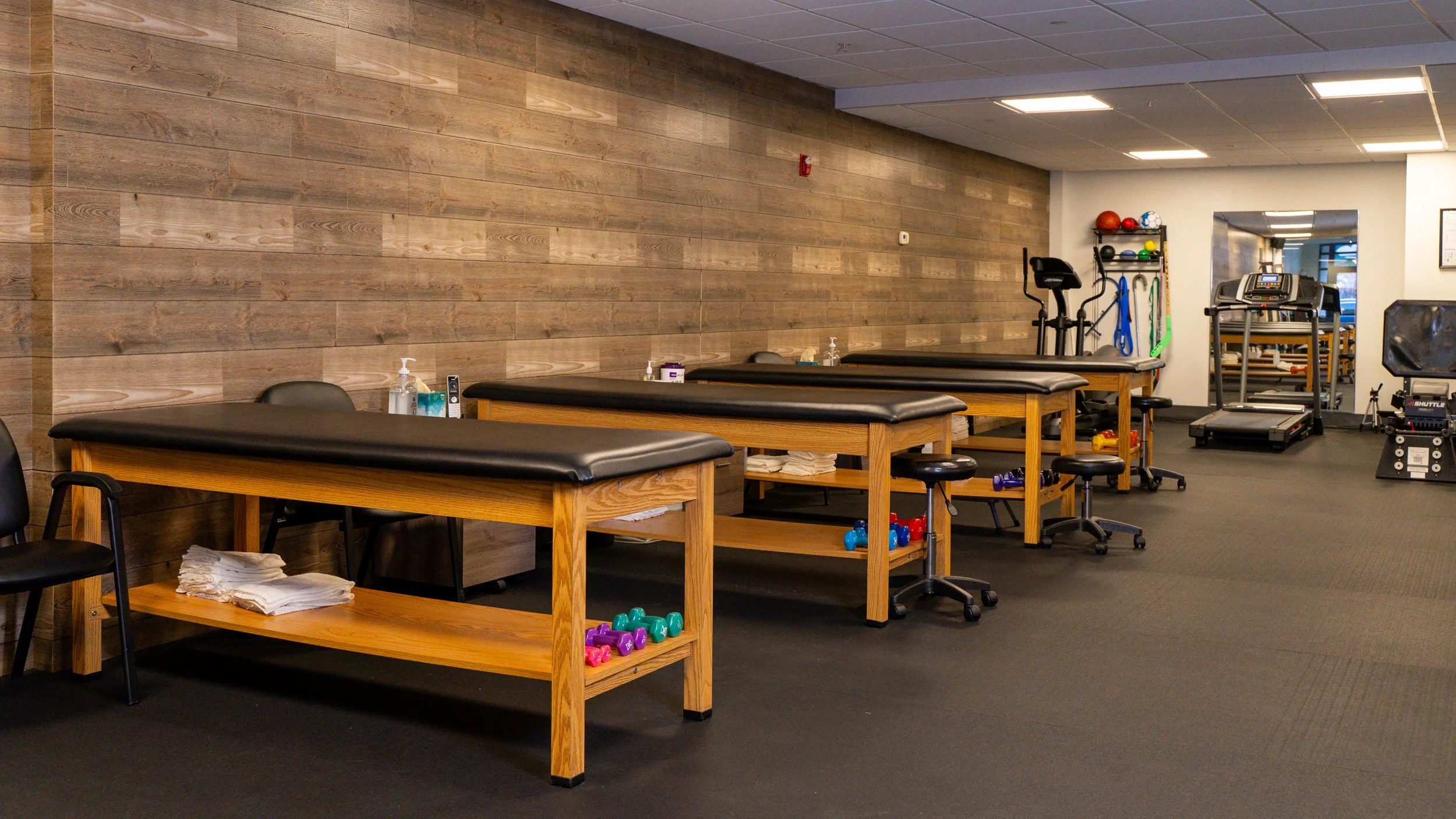 Interior of a physical therapy clinic with wooden treatment tables, chairs, hand weights, and exercise equipment like a treadmill and exercise balls