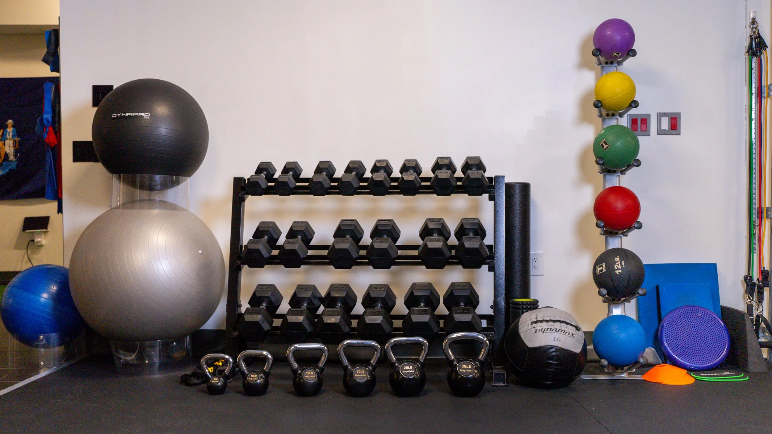 Gym equipment including dumbbells, kettlebells, exercise balls, medicine balls, and resistance bands organized against a wall.