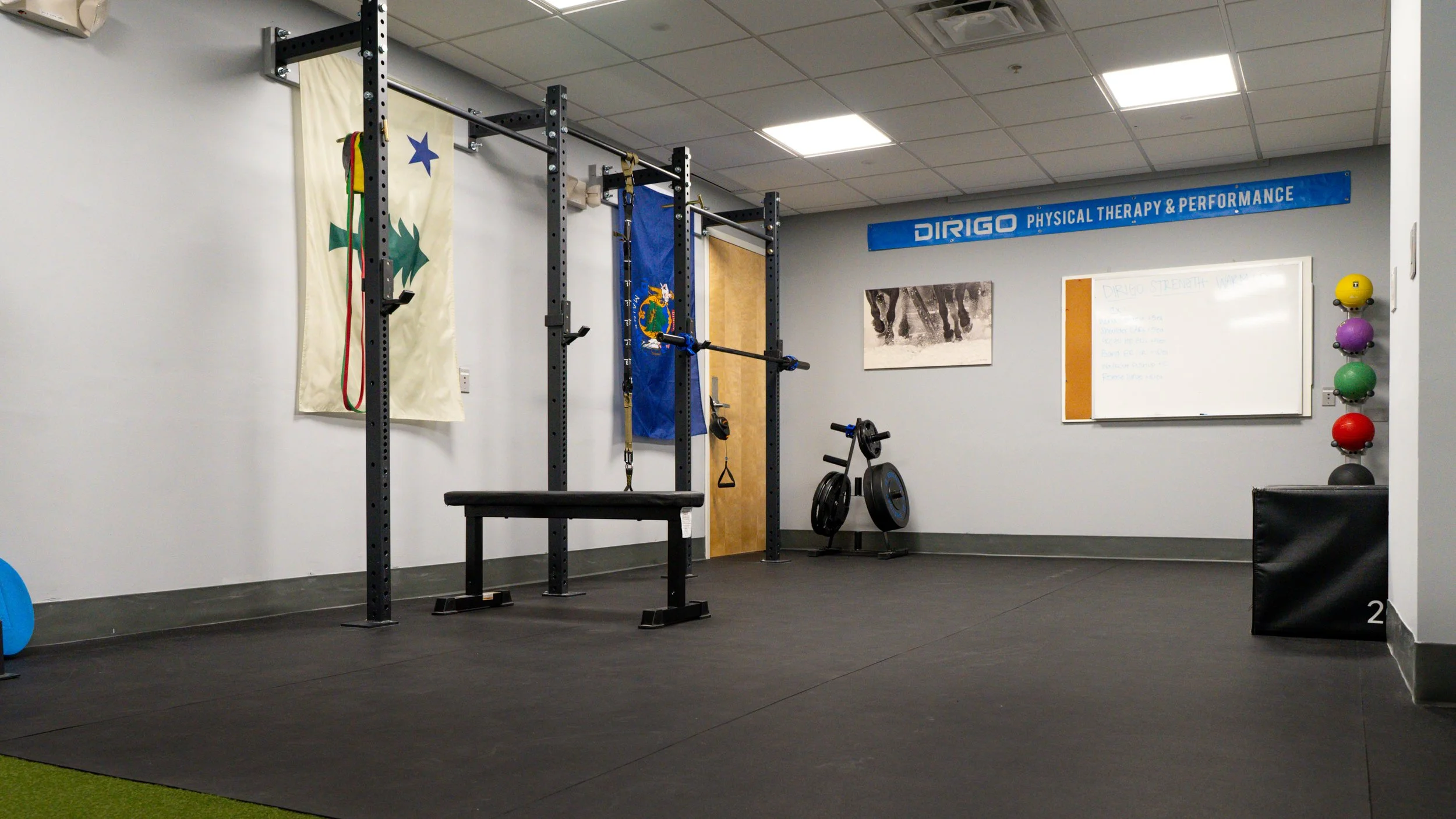 Gym room with exercise equipment, including a weight rack, bench, stationary bike, wall decor, and a whiteboard with workout notes.