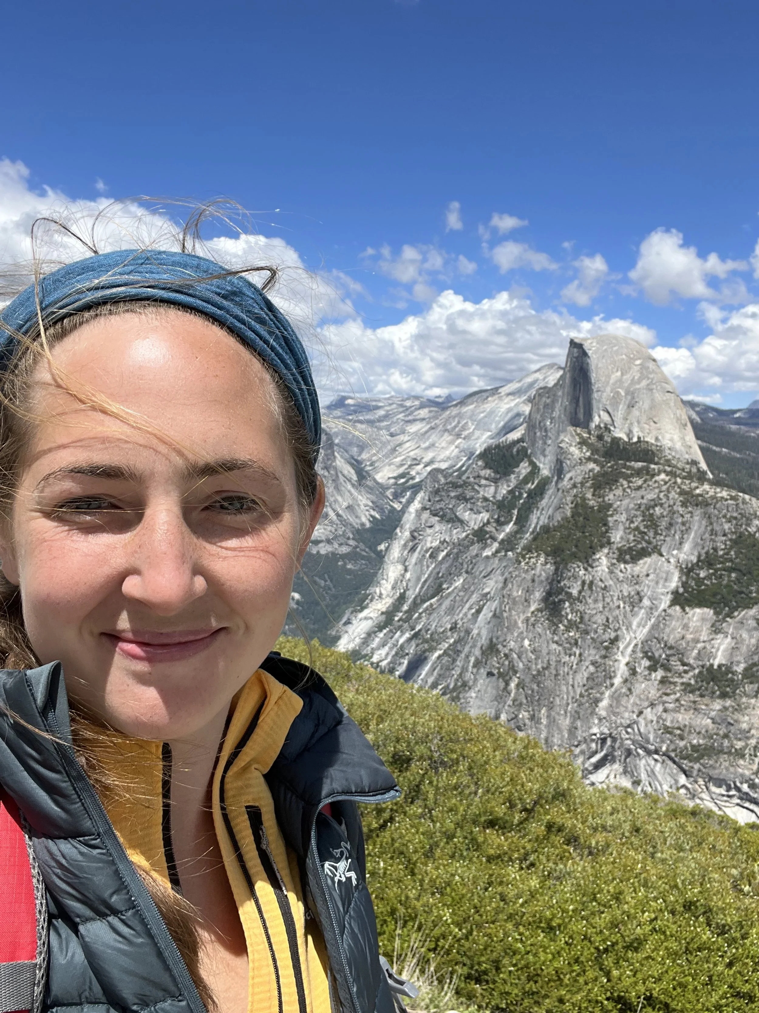 Woman smiling in front of a scenic mountain view with Half Dome in the background under a blue sky.