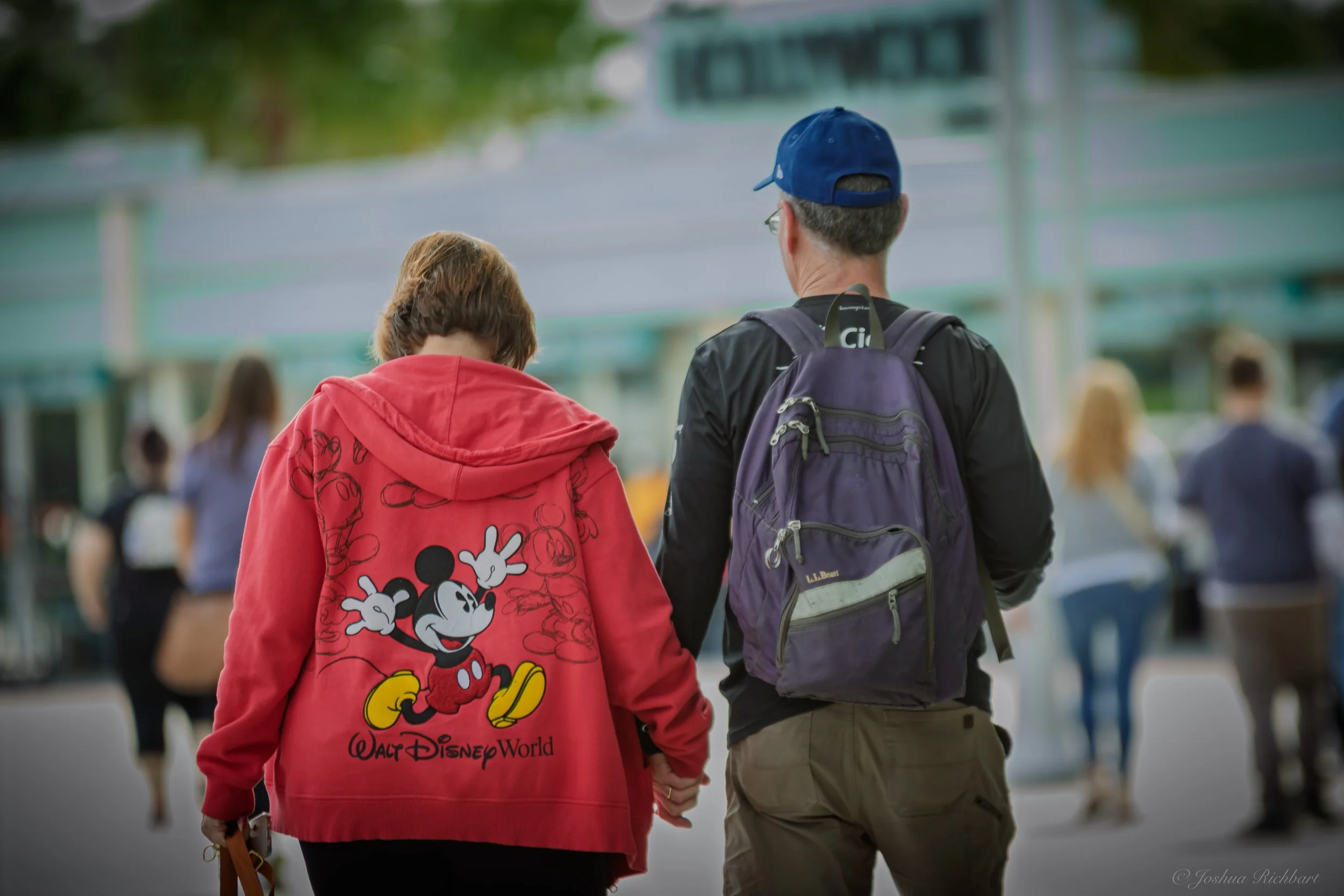 A woman wearing a red Mickey Mouse jacket and a man with a purple backpack walking outdoors at Walt Disney World, with other people in the background.