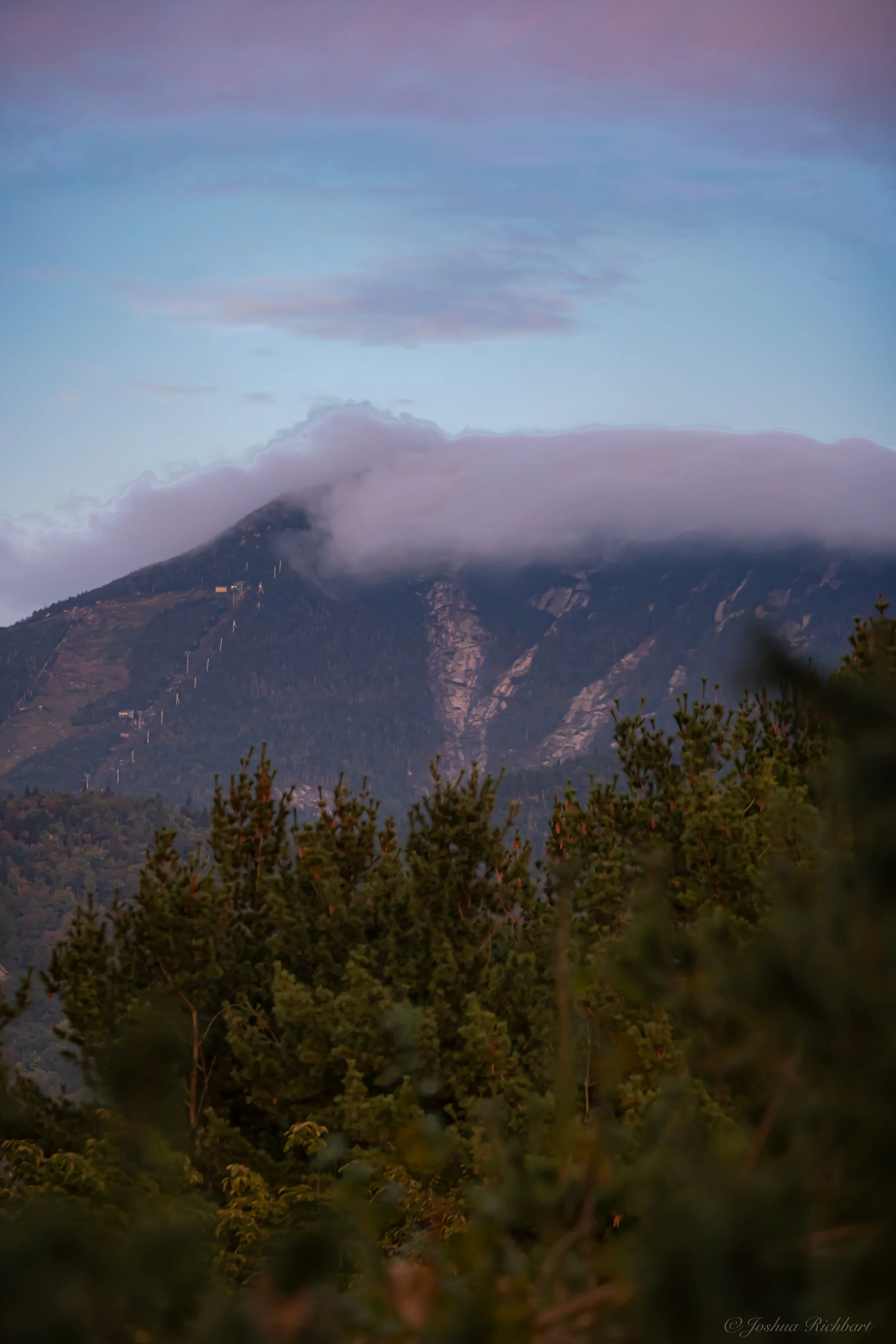 Whiteface mountain partly covered by clouds with green trees in the foreground.