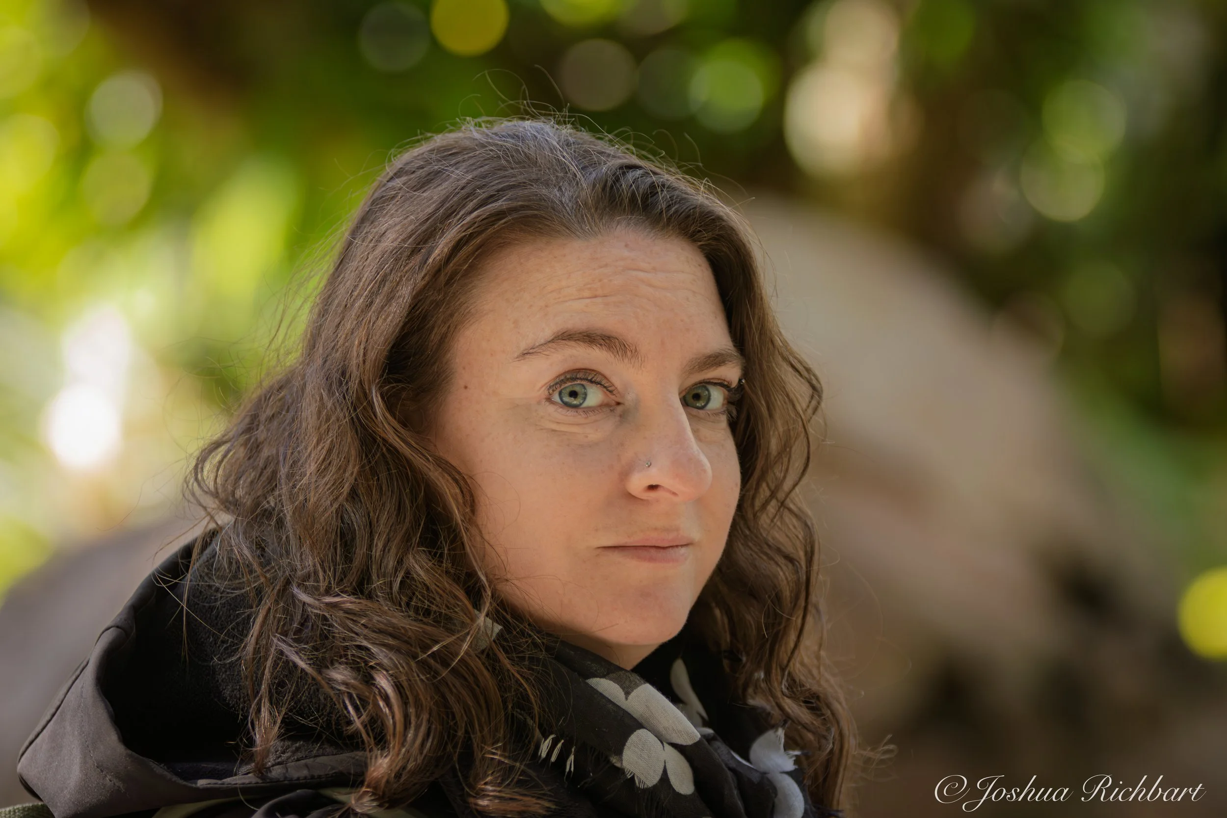 Close-up of a woman with curly brown hair and blue eyes, wearing a black and white scarf, outdoors with a background of blurred green and yellow foliage.