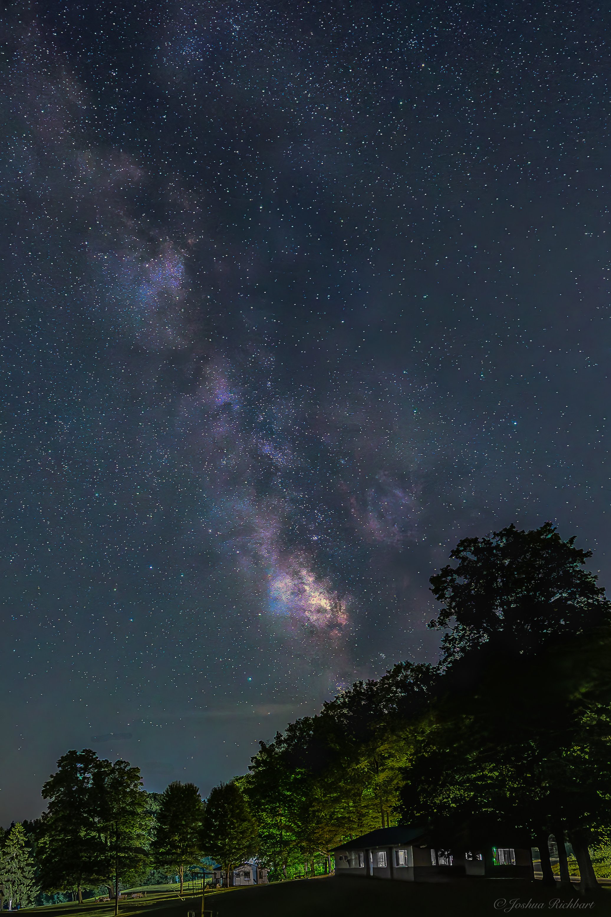 Night sky filled with stars and the Milky Way galaxy, with a silhouette of trees and a house at the bottom of the image.