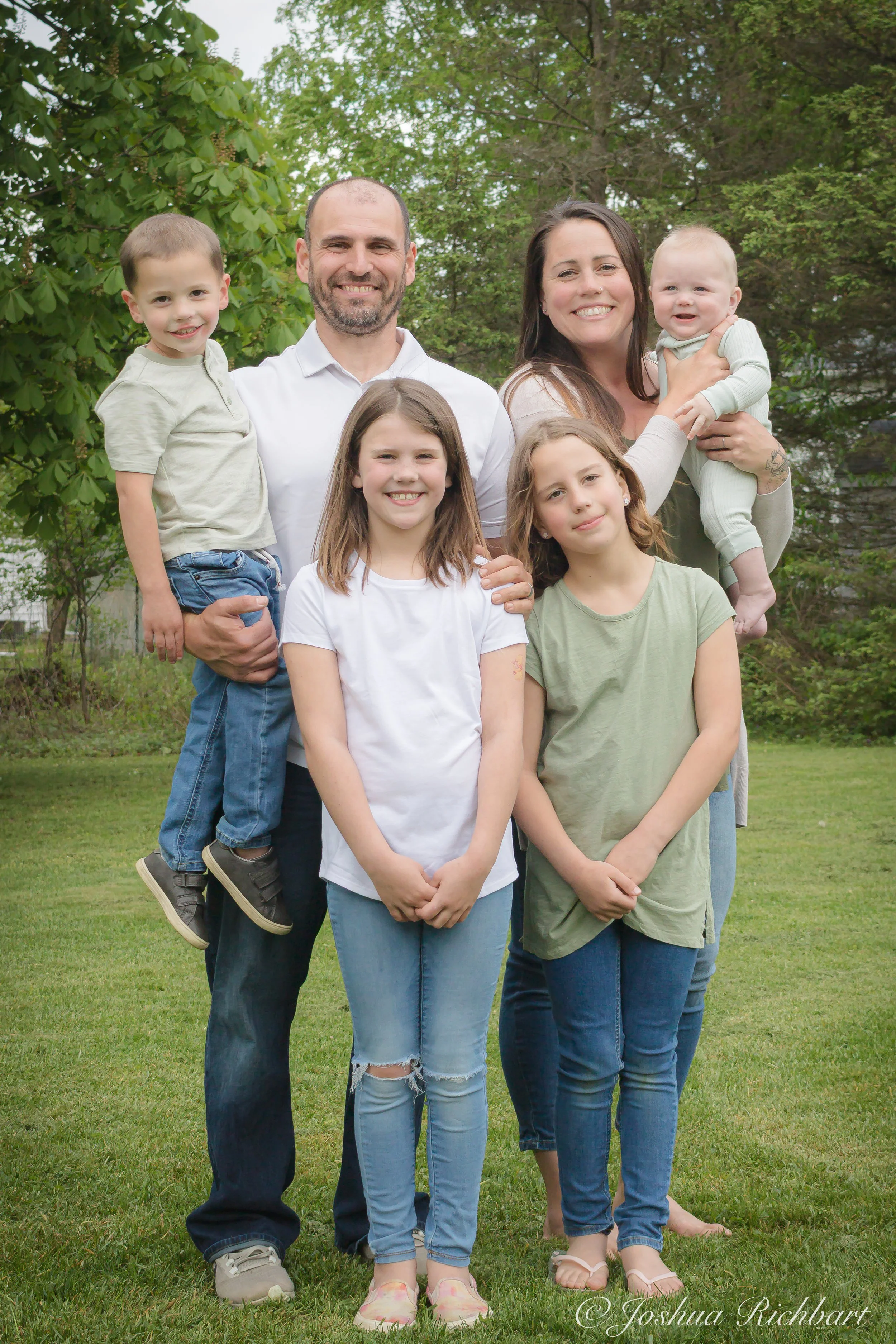 A family of seven standing outdoors on a grassy lawn, surrounded by green trees. Two adults, a man and a woman, are in the back, with four children in front. The man is holding a young boy, and the woman is holding a baby. The children are smiling, a