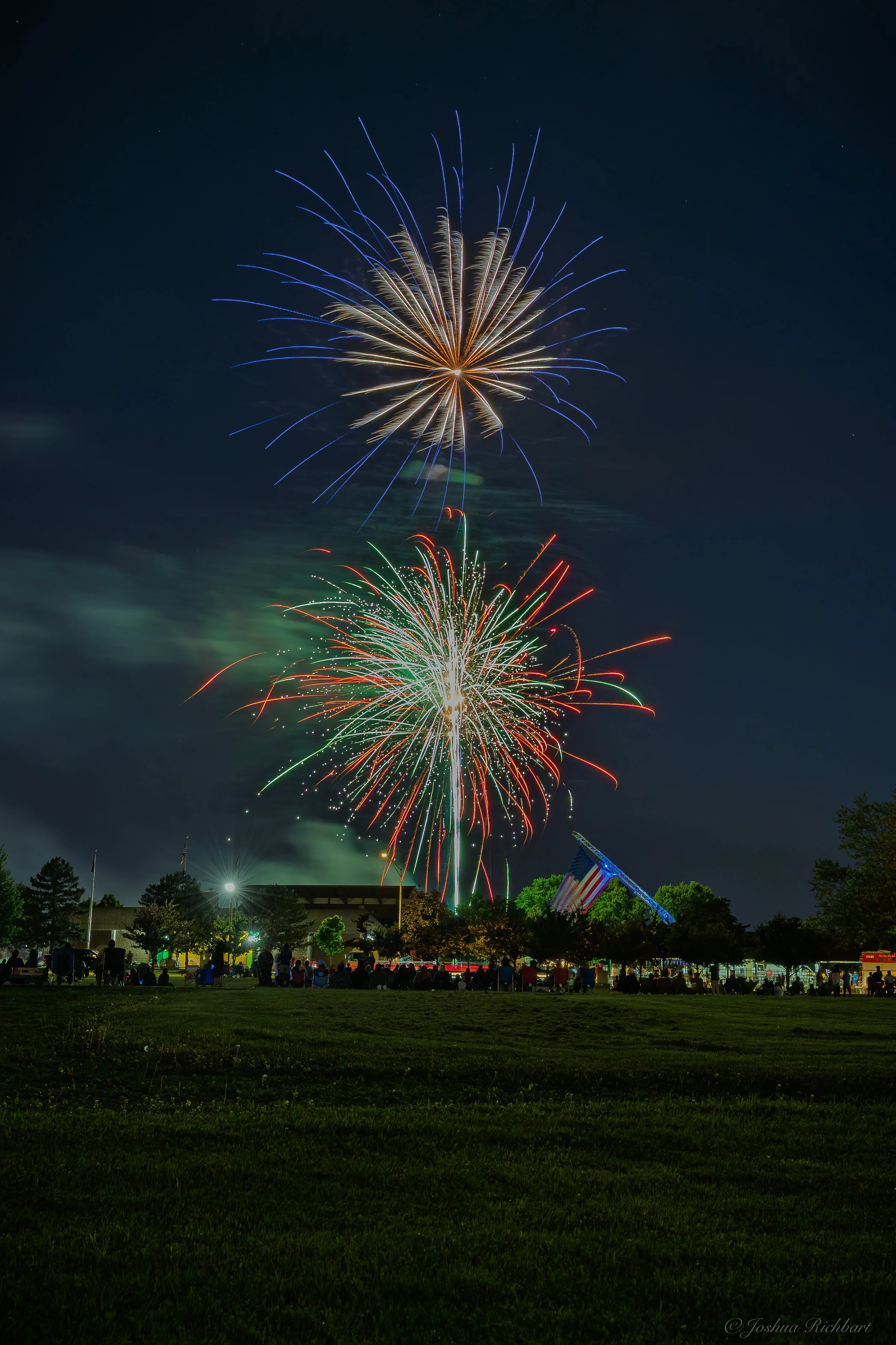 Fireworks display lighting up the night sky over a park with silhouetted spectators, trees, and flags.