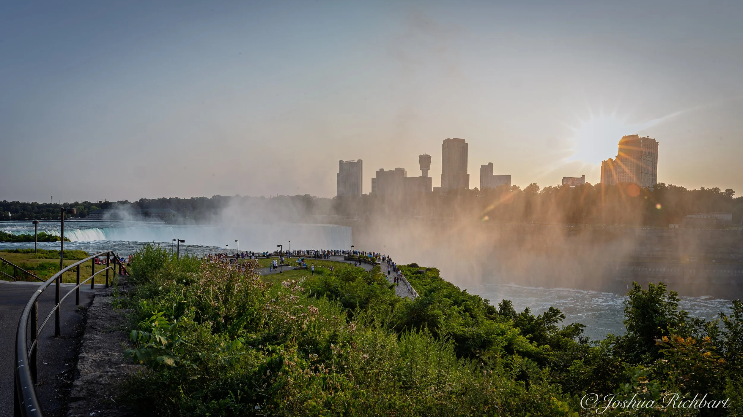 View of Niagara Falls at sunset with a crowd of tourists on the observation area, city skyline in the background, and mist rising from the falls.