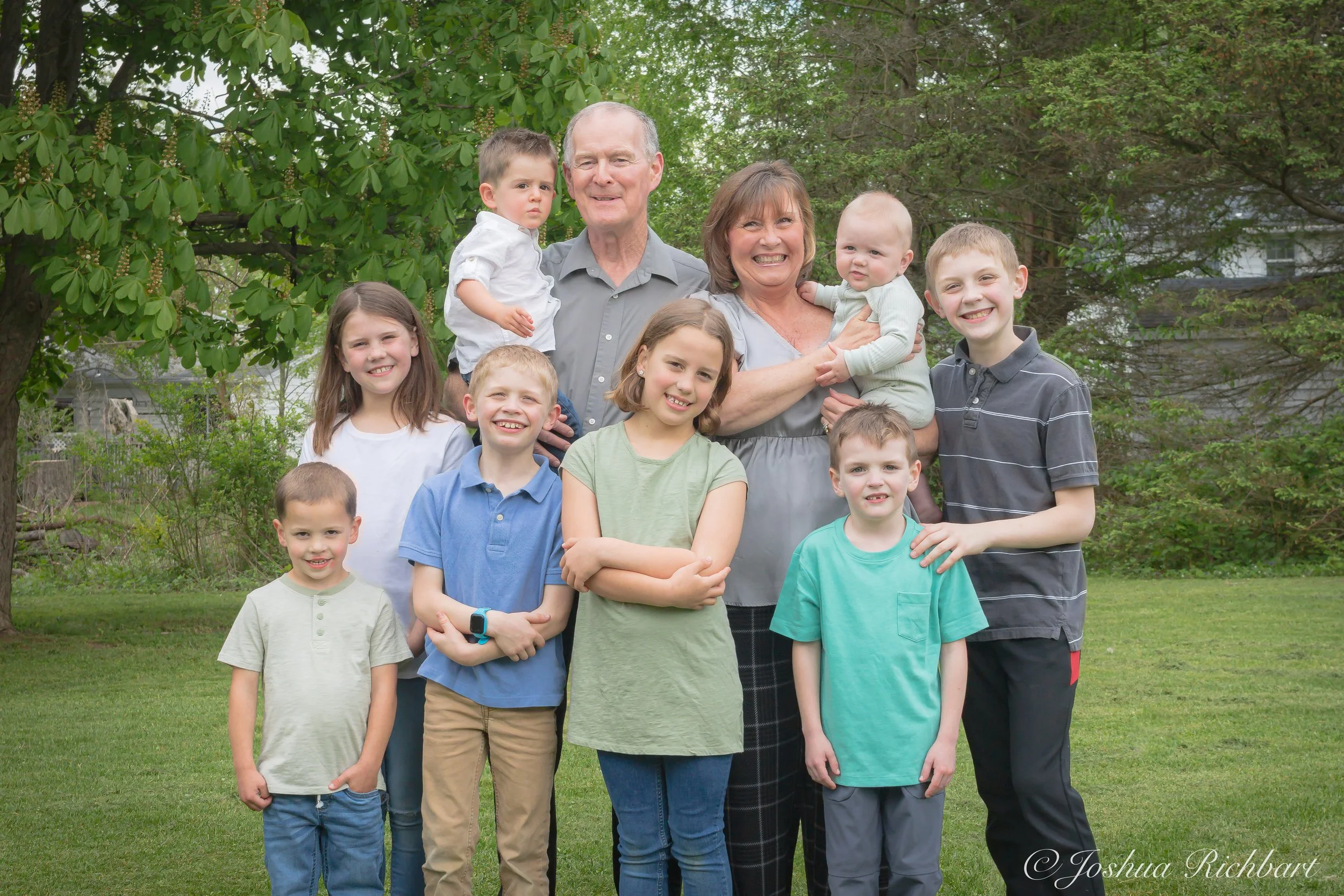 A family of eleven people, including grandparents, parents, and children, standing outdoors in a lush green yard with trees in the background, smiling at the camera.