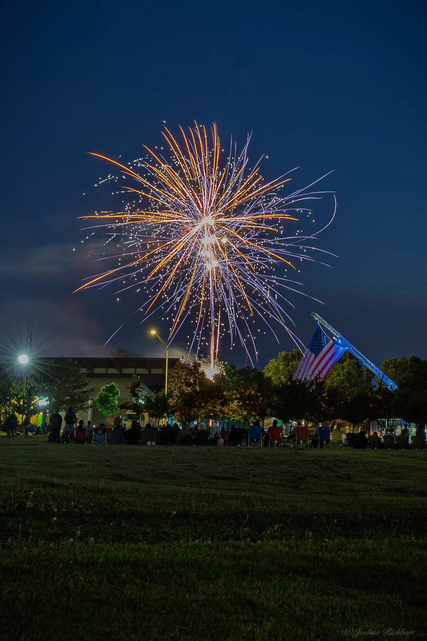 Fireworks light up the night sky during a celebration, with people sitting on the grass watching, and an American flag displayed on a structure.
