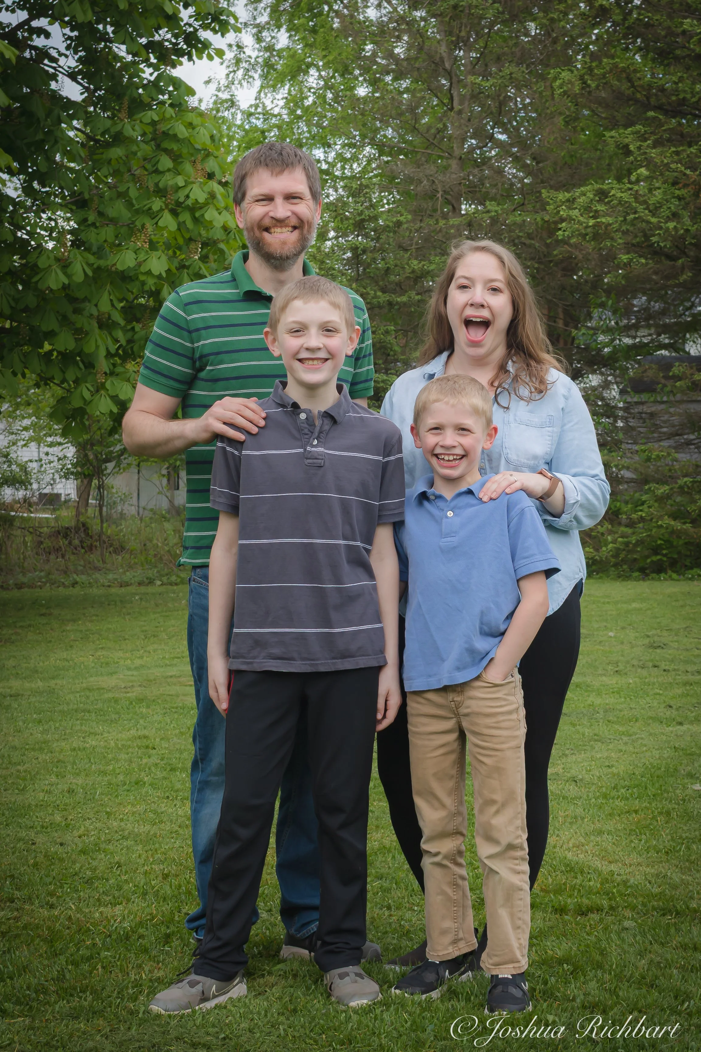 A family of four standing outdoors on a grassy lawn, smiling and laughing. The father and mother are standing behind two boys, with trees and greenery in the background.