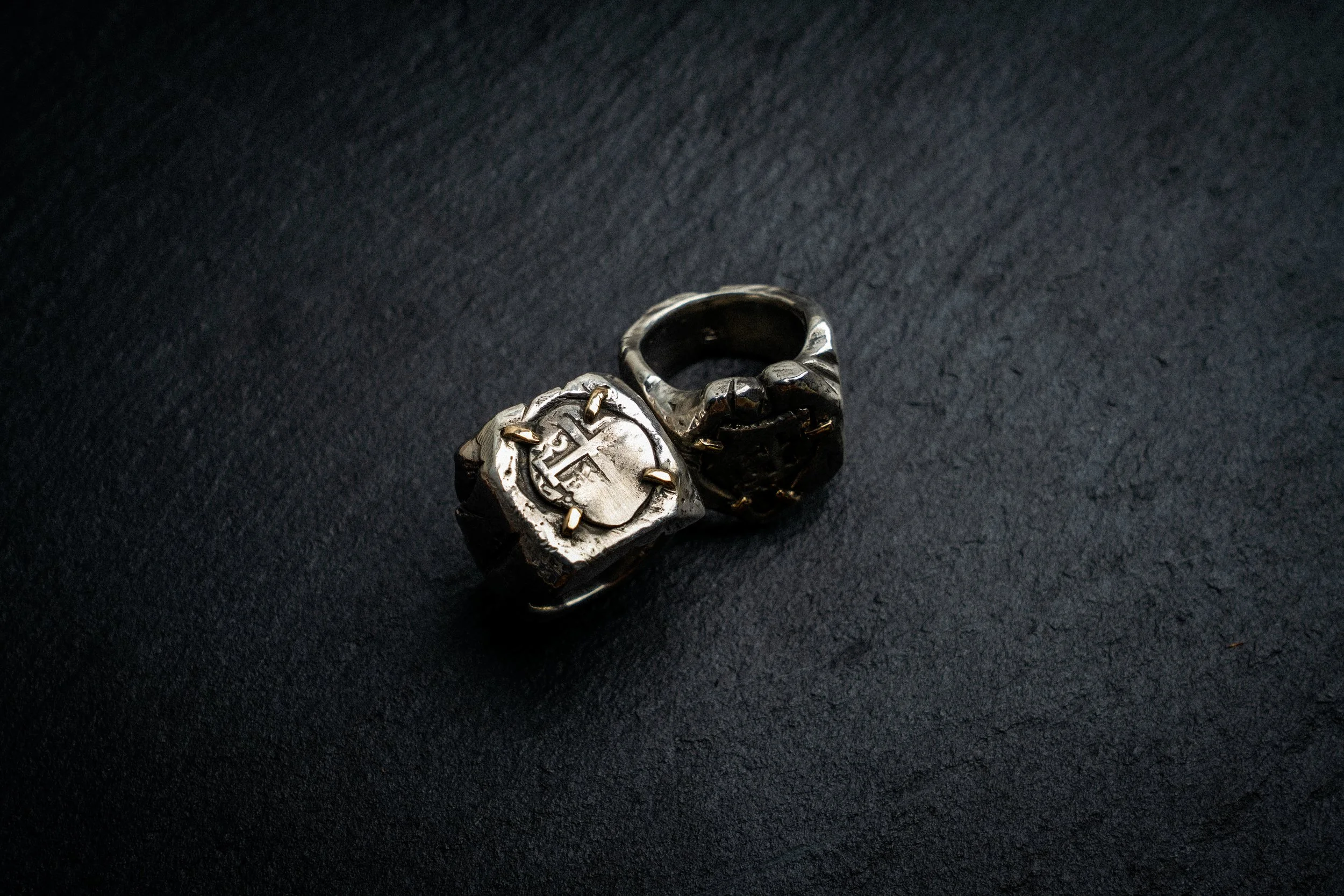 Close-up of a silver ring with a cross engraved on a heart-shaped face, resting on a black textured surface.