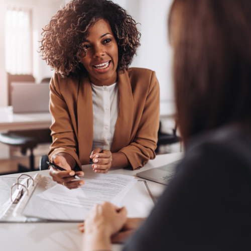 Two women having a conversation in an office, one woman with curly hair and wearing a tan blazer, smiling and holding a pen, the other woman blurred in the foreground.