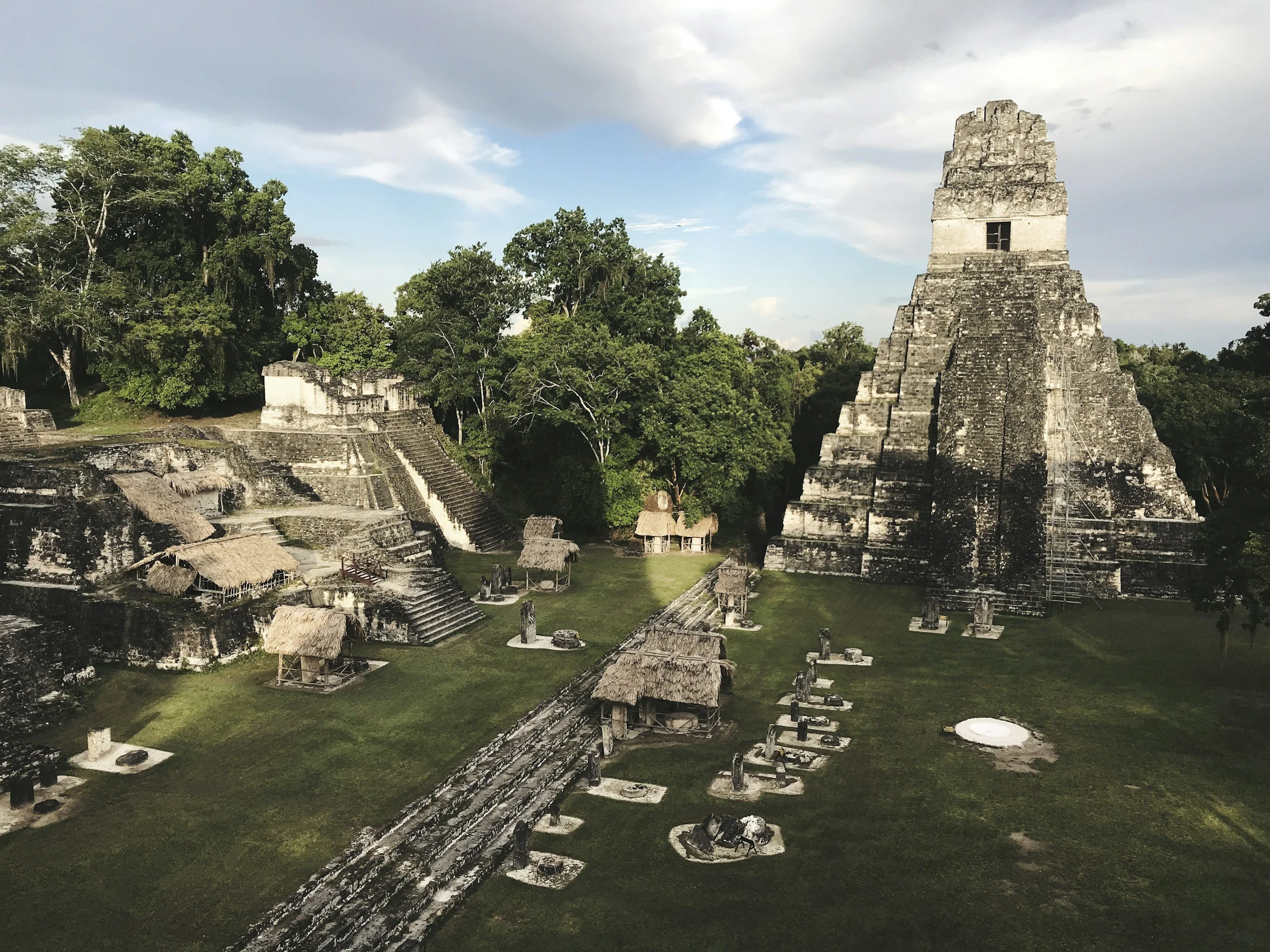 Ancient Mayan pyramid surrounded by jungle, with smaller stone structures and thatched huts in front.
