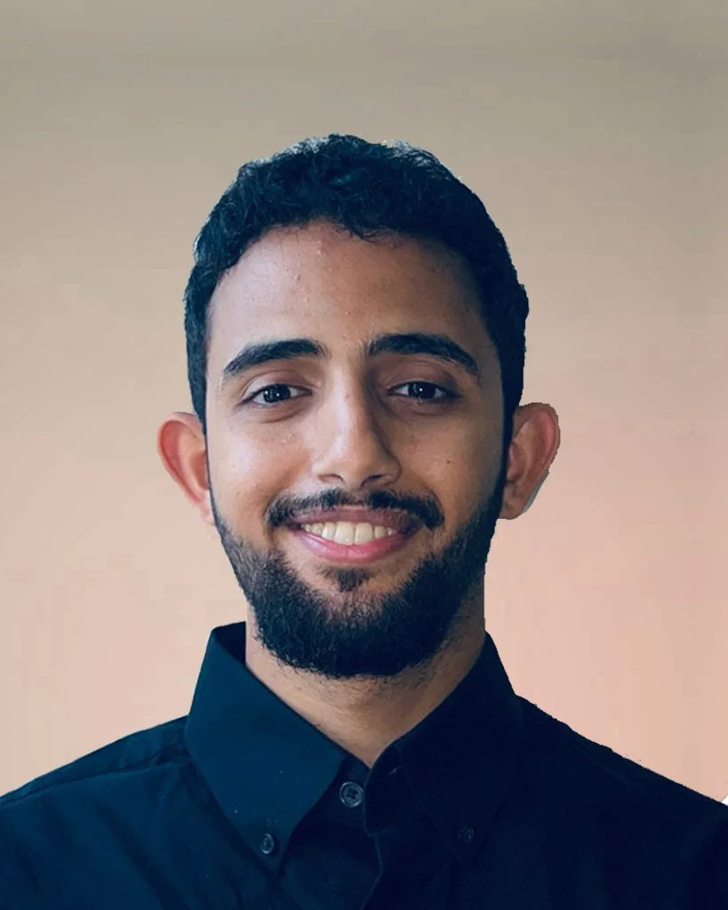 A young man with dark curly hair, a beard, smiling, wearing a black collared shirt, against a plain beige background.