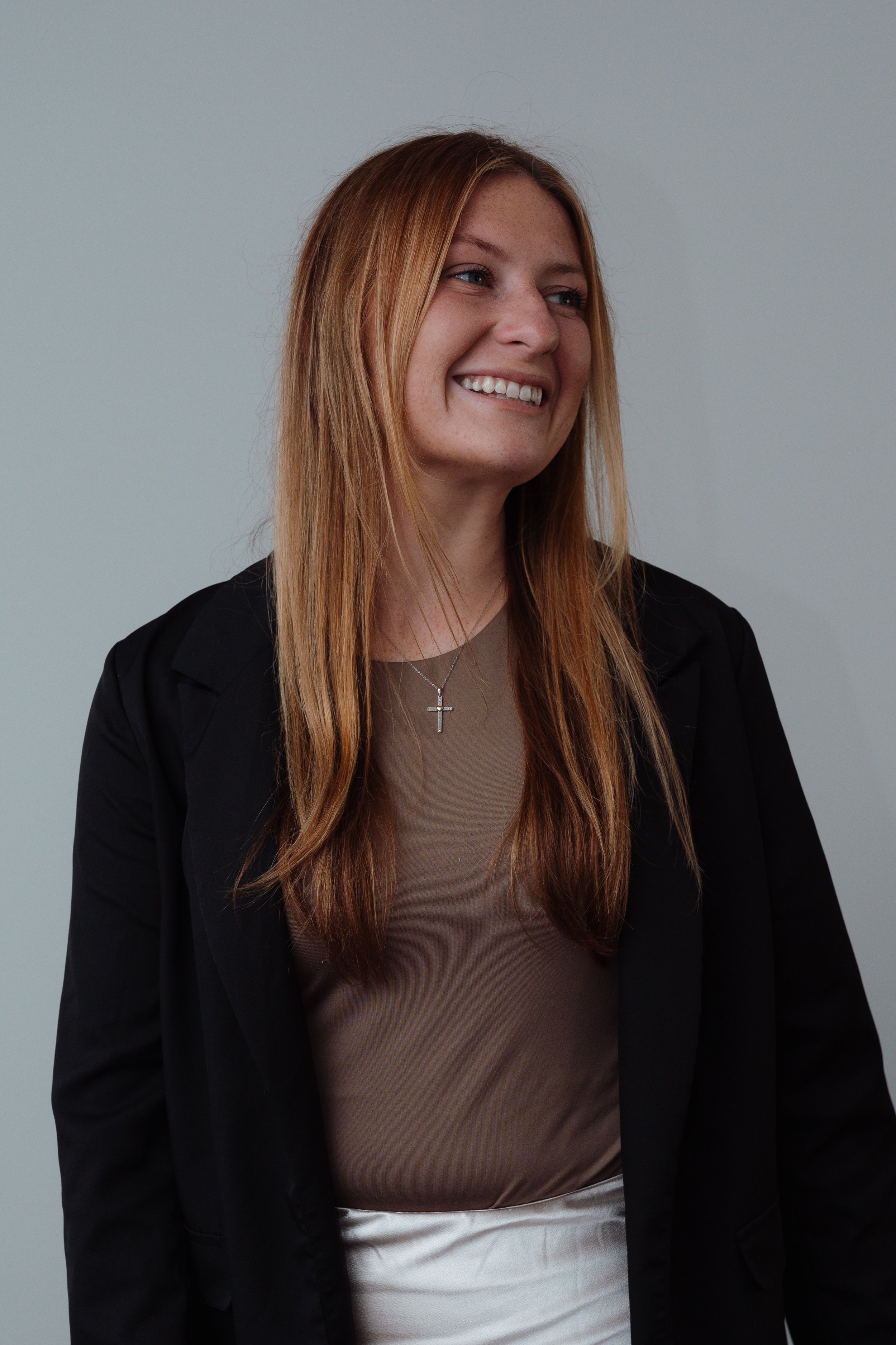 A young woman with long red hair wearing a black blazer, taupe top, and white skirt, smiling and looking to the side against a plain background.