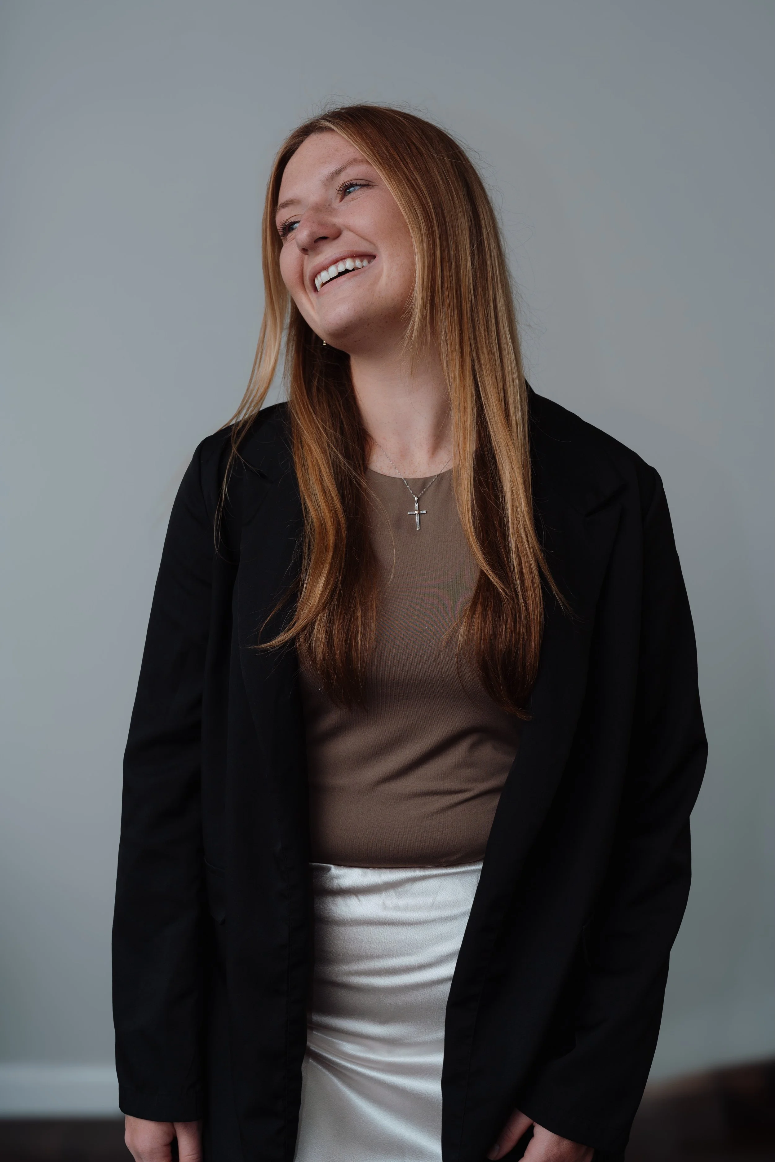 A woman with long red hair smiling and looking to her left, wearing a black blazer, a taupe top, a white skirt, and a cross necklace, standing against a plain light grey background.