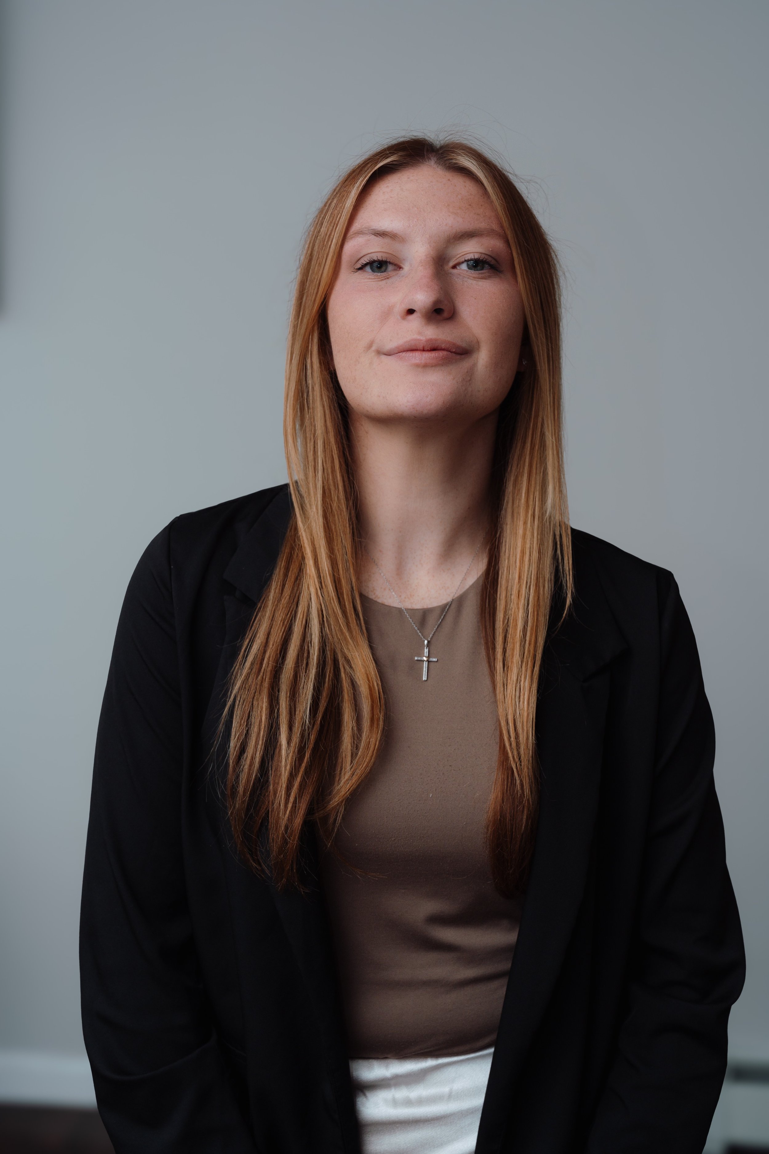 A woman with long red hair and light skin, wearing a black blazer, a taupe top, and a cross necklace, looking at the camera with a slight smile, standing in front of a plain gray background.