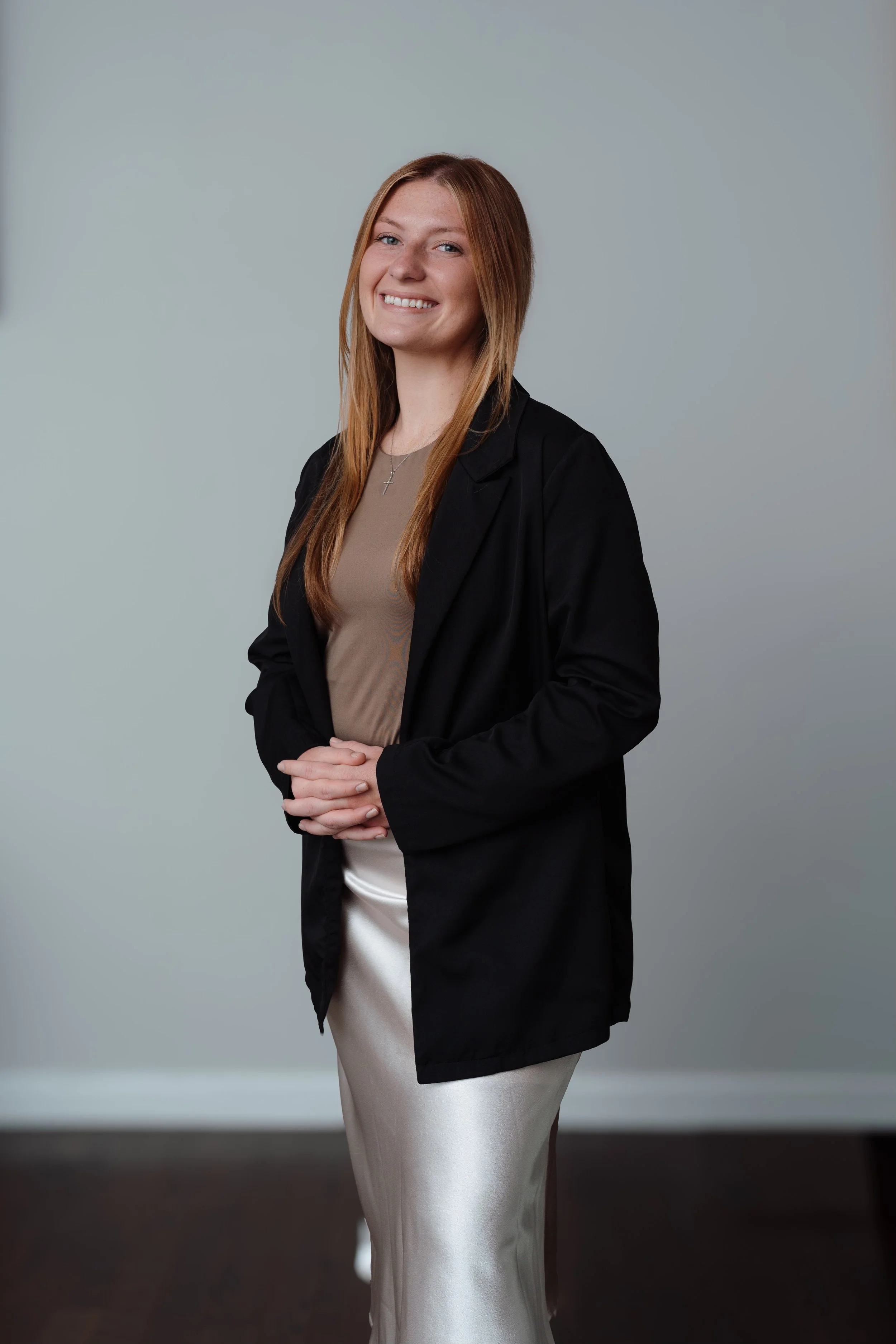 A woman with long red hair smiling, wearing a black blazer over a beige top and a cream-colored dress, standing against a plain light gray background.