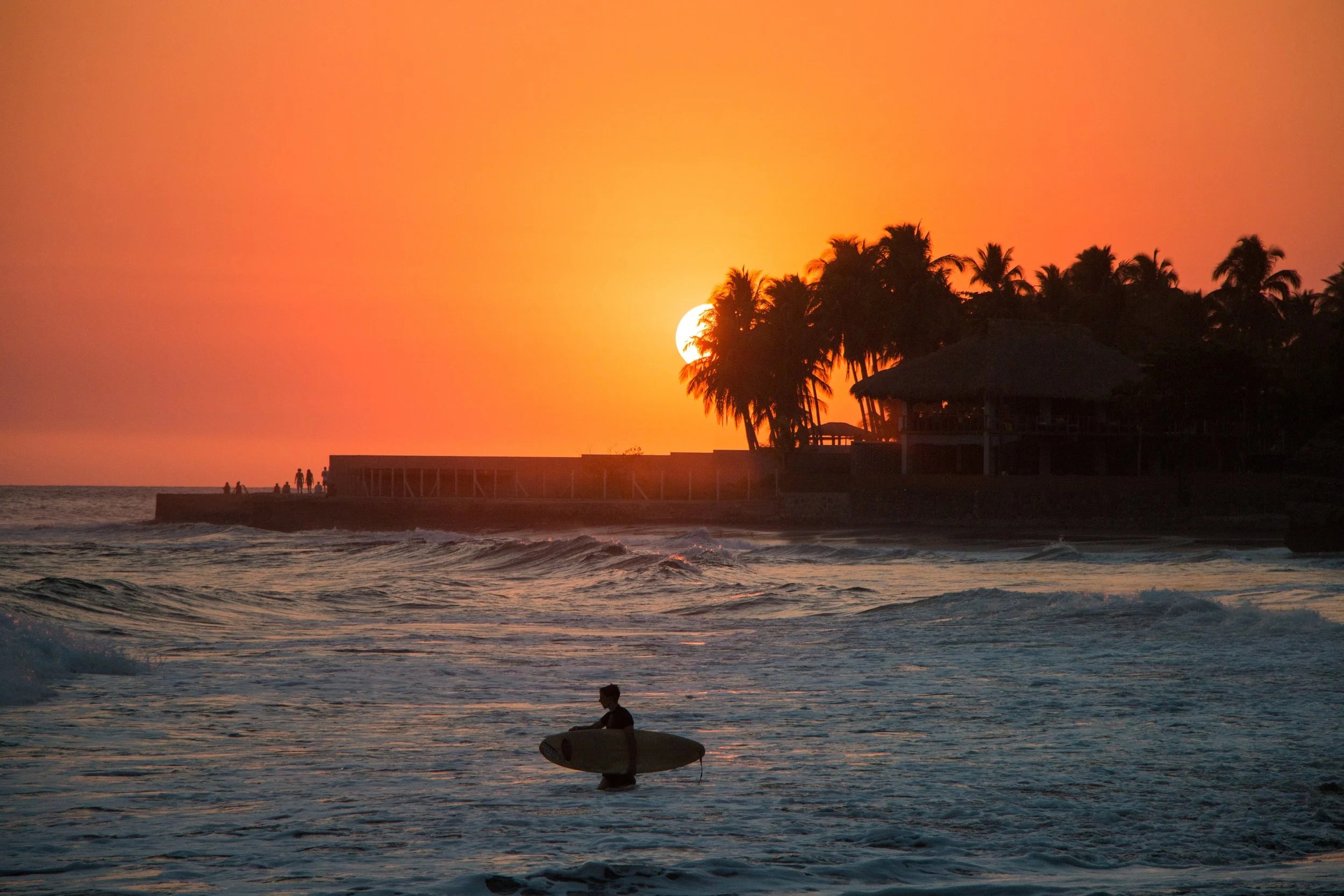 A person holding a surfboard standing in the ocean at sunset, with palm trees and a building in the background.