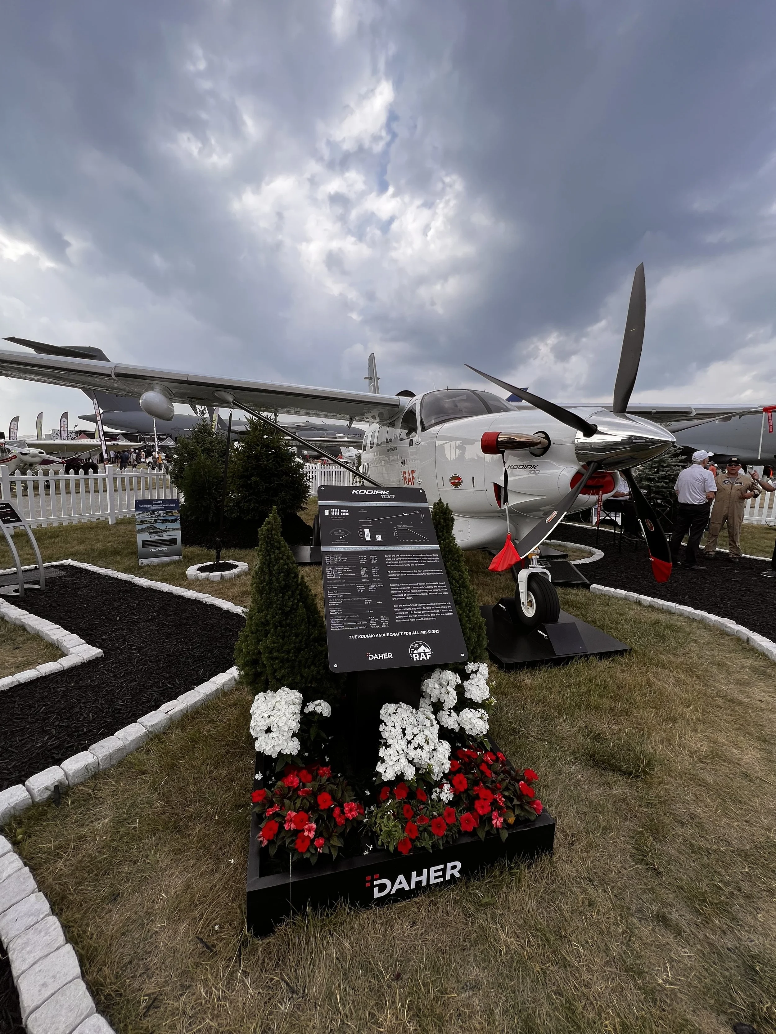 A small aircraft called KODIAK 100 displayed outdoors at an airshow, surrounded by flowers and informational signage, with a partly cloudy sky overhead.