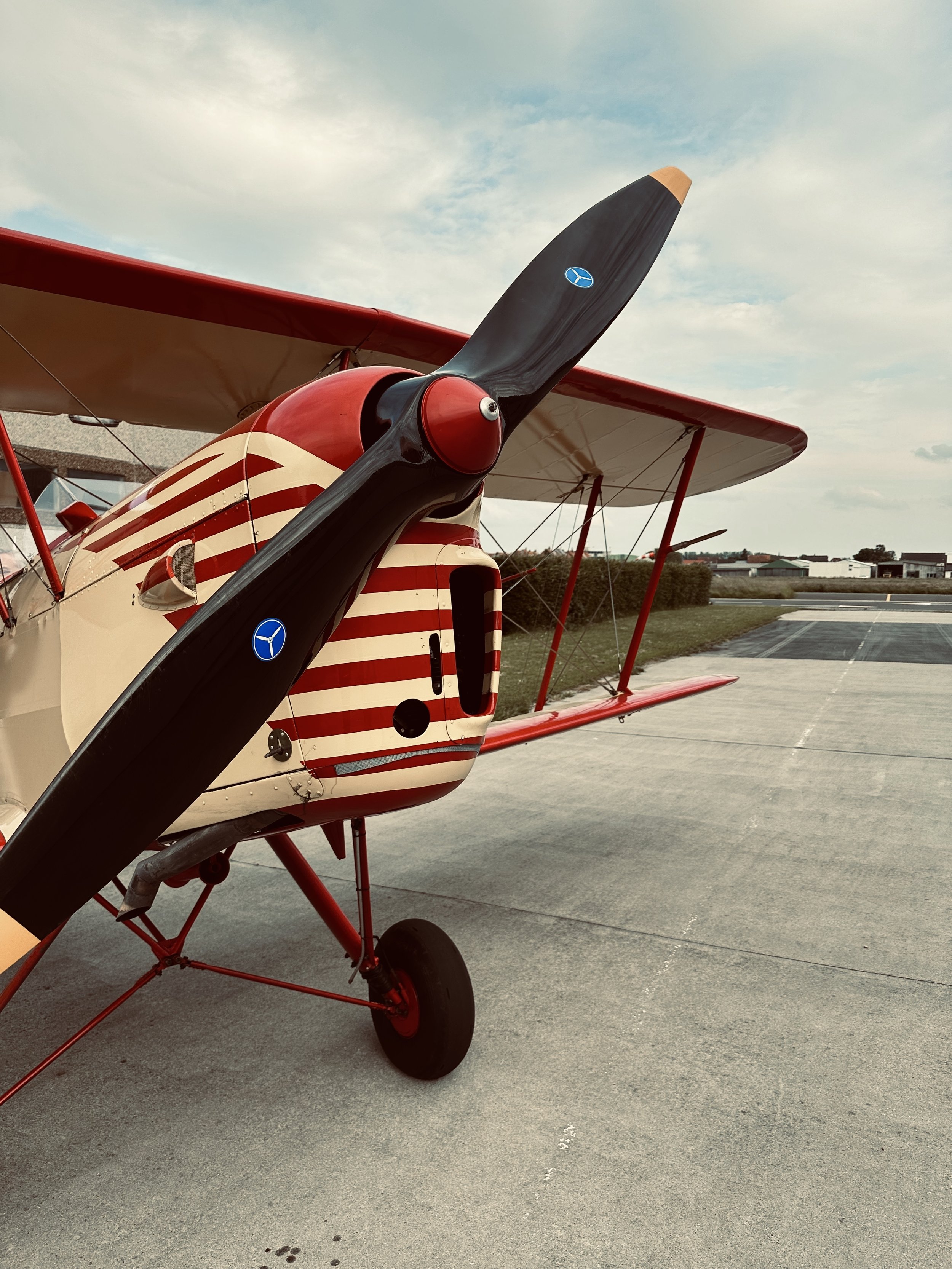 A small red and white vintage biplane with a striped pattern on the fuselage, sitting on a tarmac runway under a partly cloudy sky.