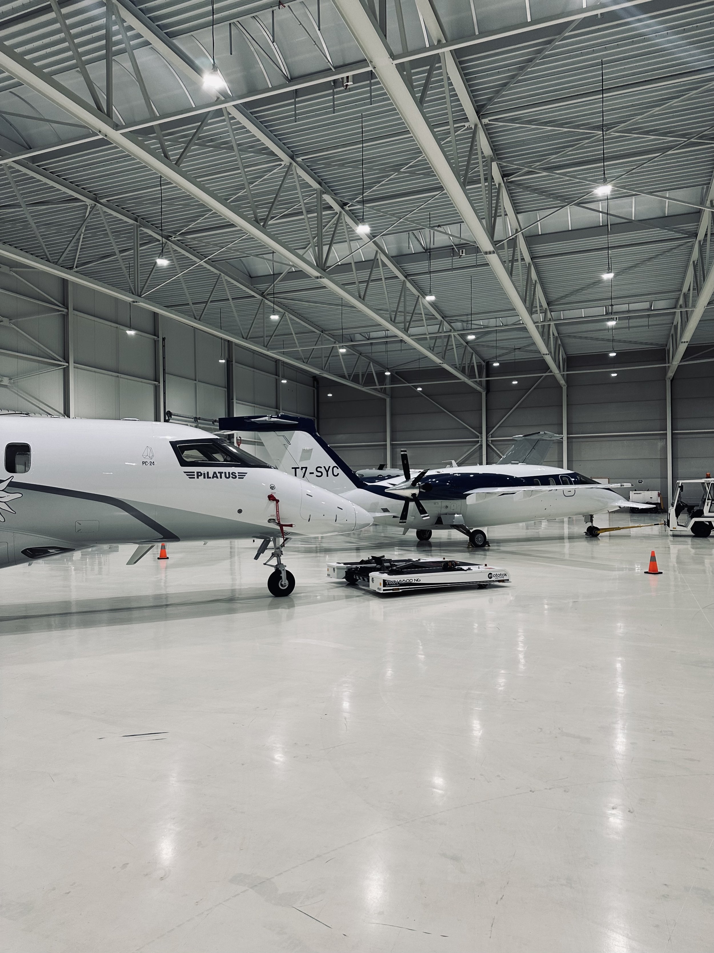 Interior of an airplane hangar with two small private airplanes parked inside. The hangar has a high metal ceiling with industrial lighting, and the floor is clean and shiny. There are cones and maintenance equipment around the planes.
