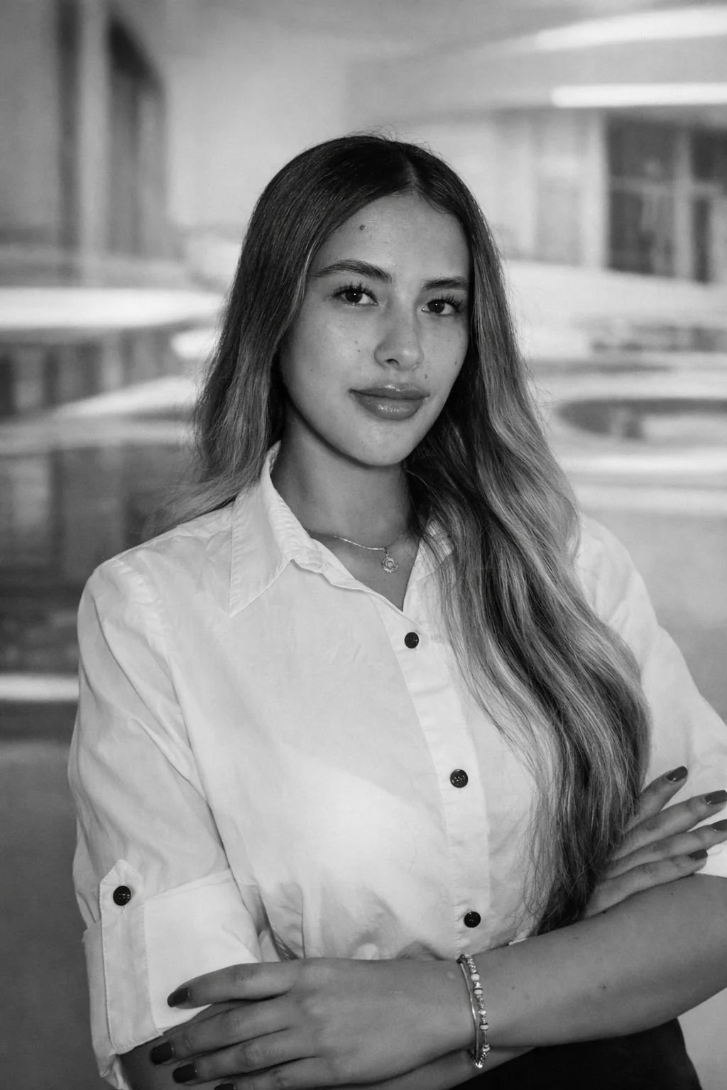 Retrato de una mujer joven con cabello largo y liso, vestida con blusa blanca, con brazos cruzados y sonriendo levemente, en un fondo difuso que parece un interior moderno.