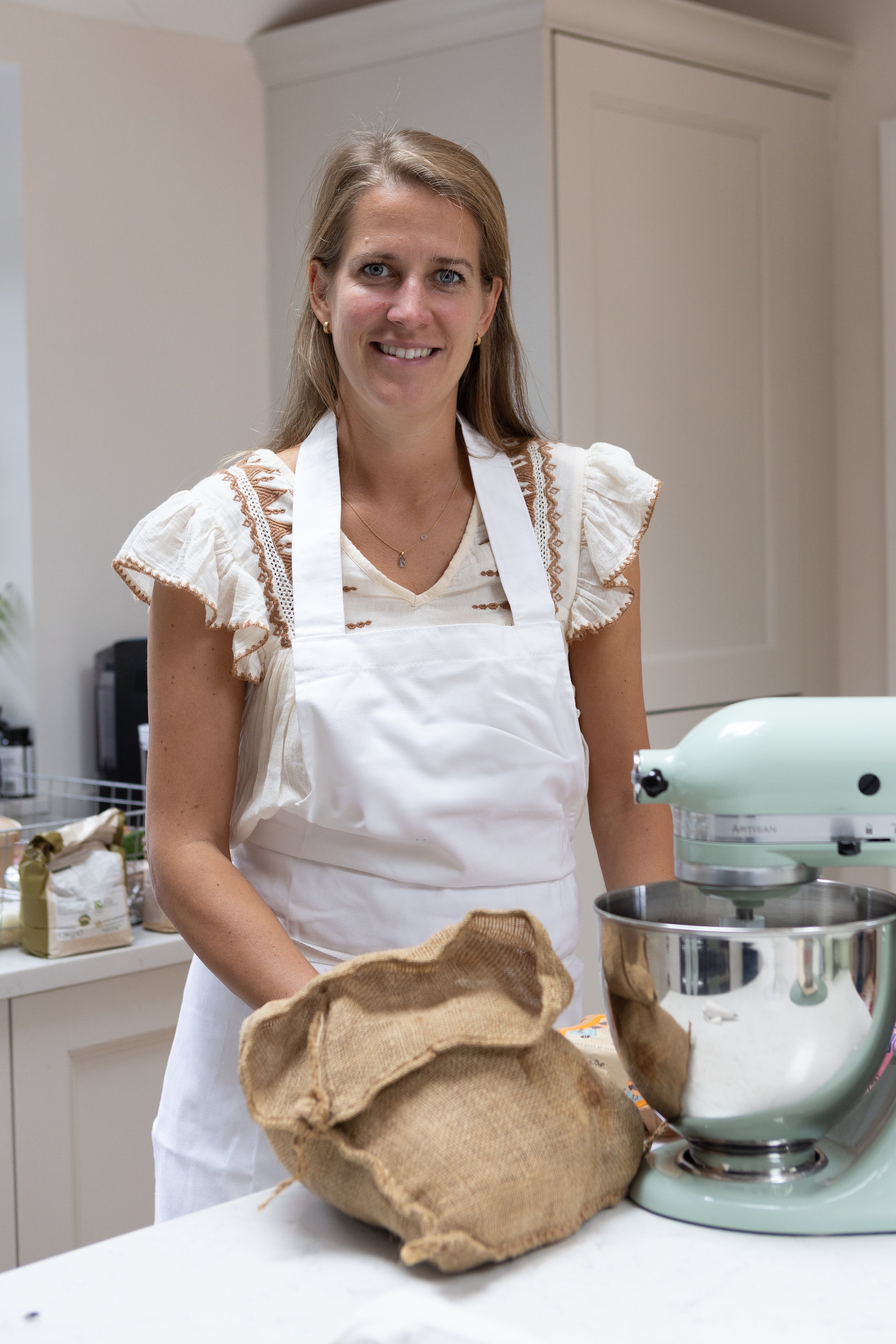 Woman in a white apron baking in a kitchen with a stand mixer and a burlap bag on the counter.