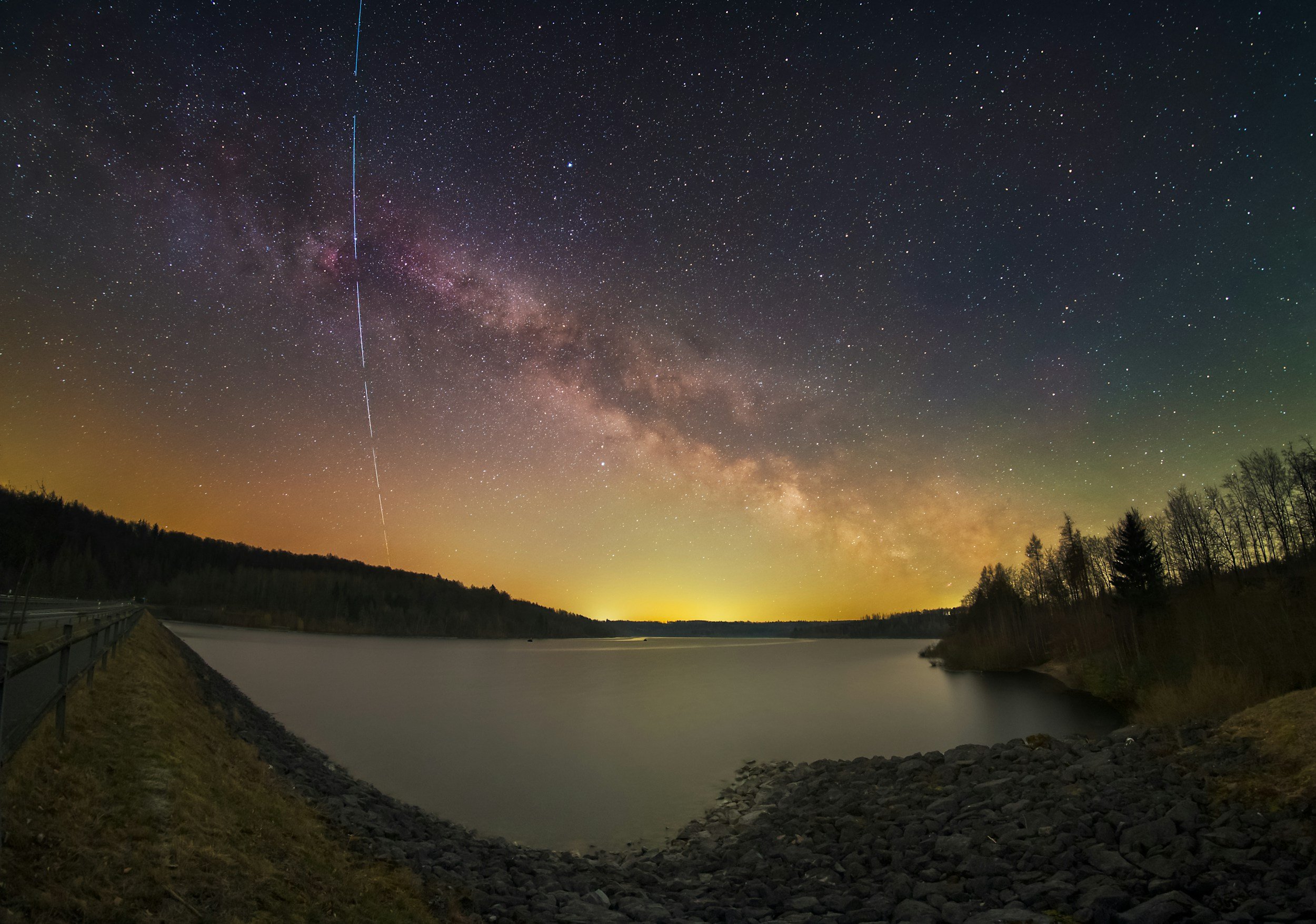 Night sky over a lake with the Milky Way galaxy and a meteor streaking across the sky, surrounded by silhouetted trees and rocky shoreline.