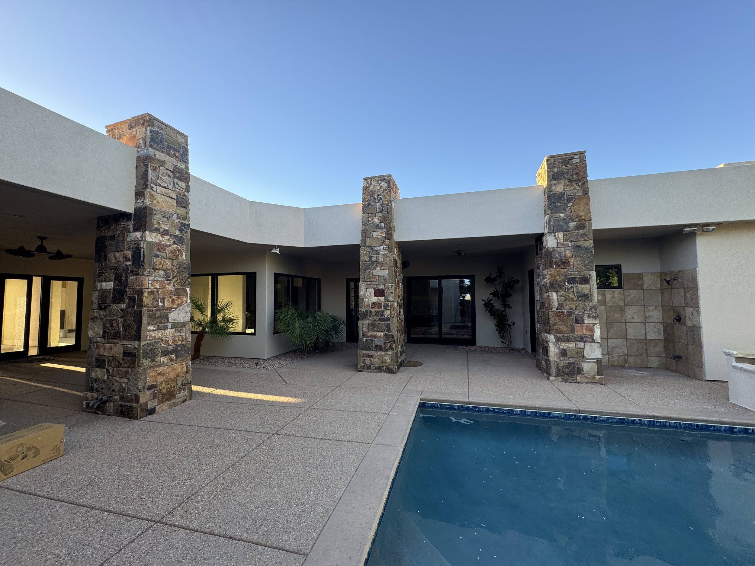 Backyard view of a modern house with stone columns, glass doors, and a pool in the foreground under a clear blue sky.