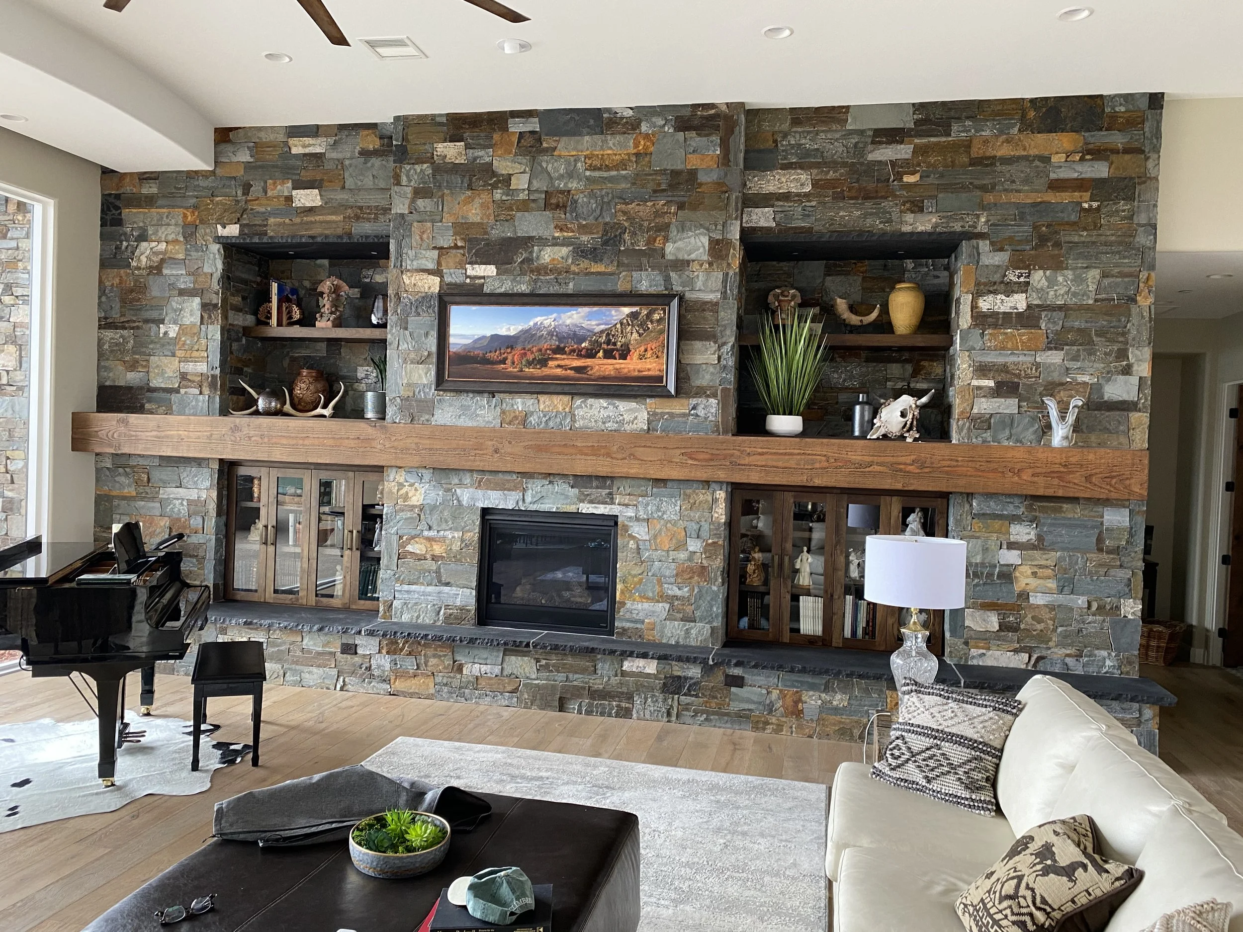 Living room with a large stone fireplace and built-in shelves, a piano, a coffee table with a bowl of plants, and a beige sofa with decorative pillows.