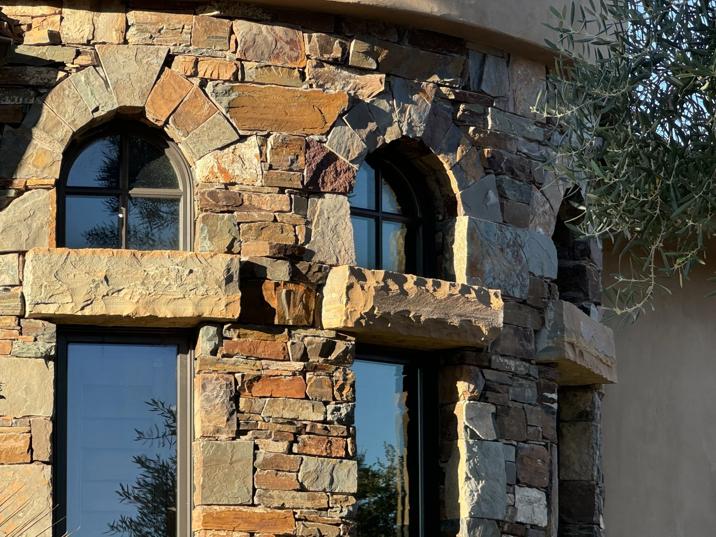 Close-up view of a stone building wall with arched window and curtain reflections, featuring various rectangular and irregular stones in natural earthy tones. Part of a tree with green leaves is visible on the right side.