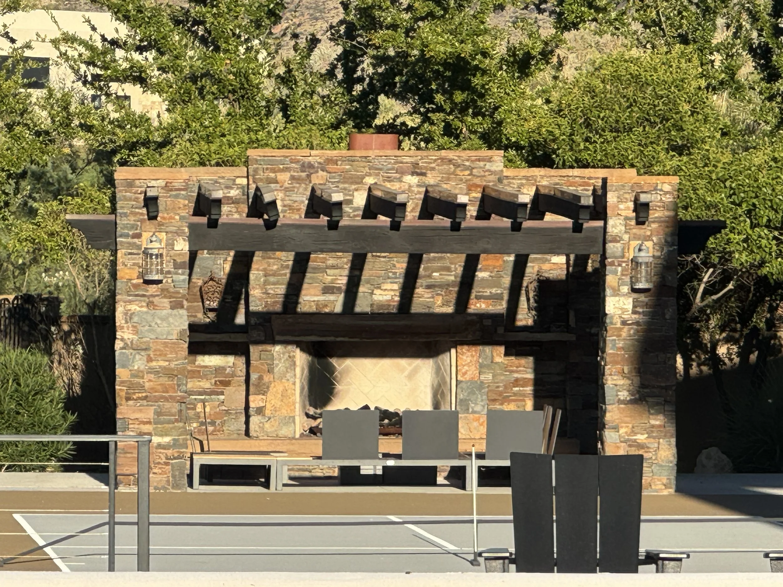 Outdoor stone fireplace with chairs and a basketball court in the foreground, surrounded by trees.